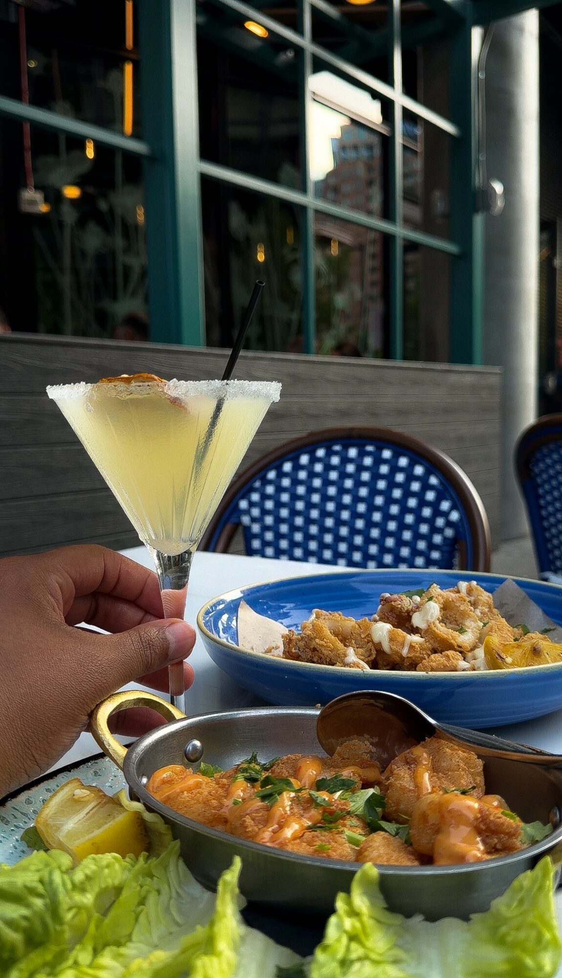 Hand holding a cocktail glass with a yellow drink, set on an outdoor table with food and blue chairs nearby.