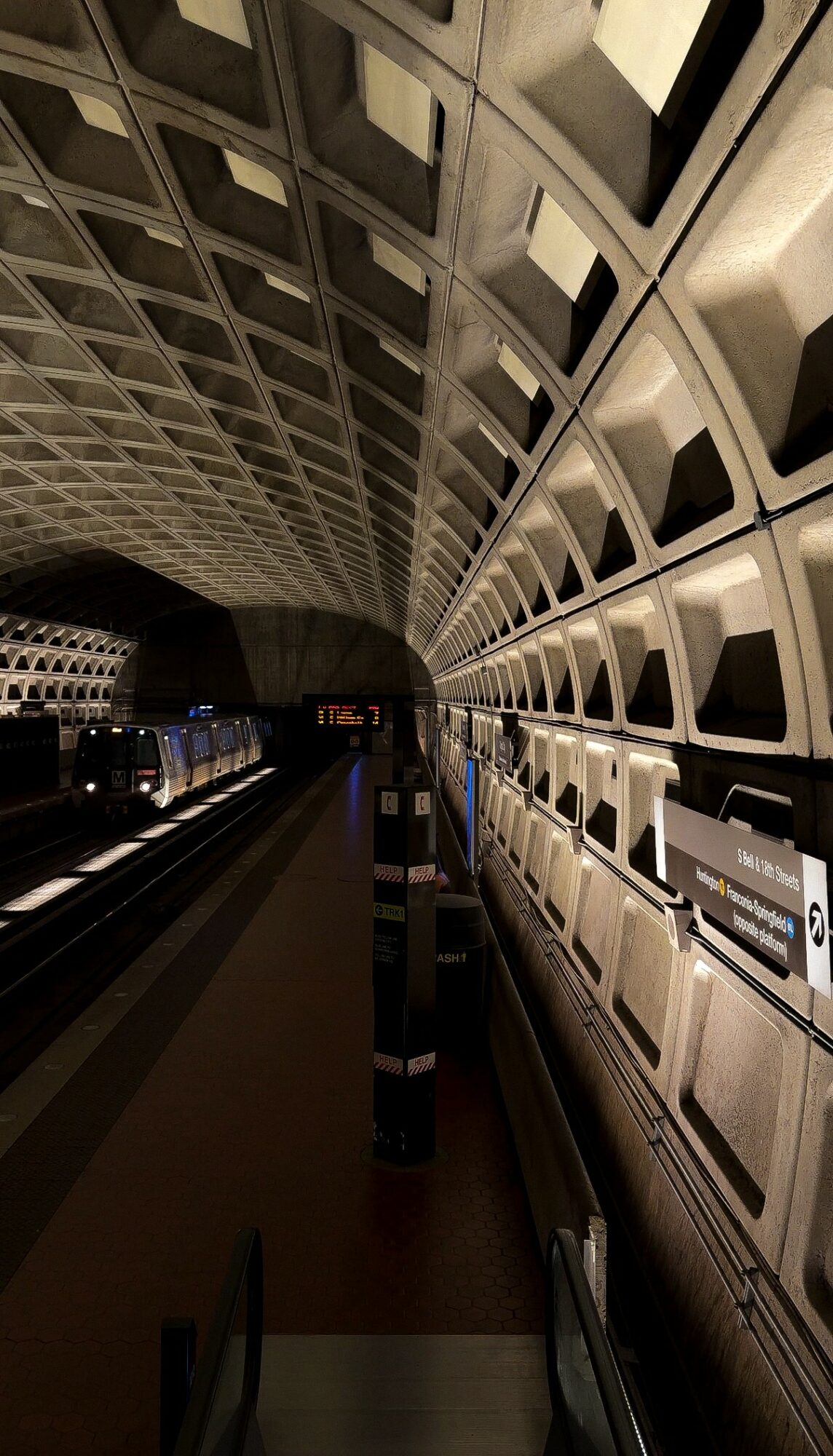 Underground station with curved wall of square panels, platform with train, and dim lighting.