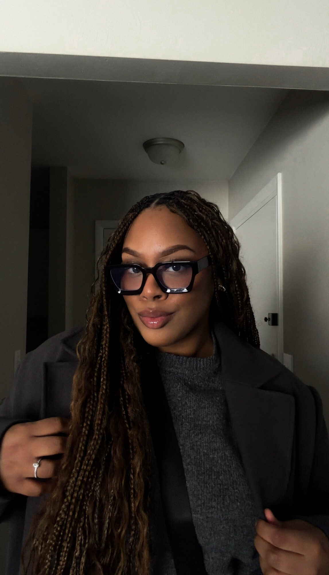 Woman with long braided hair and glasses, wearing a dark coat and gray sweater, standing indoors.