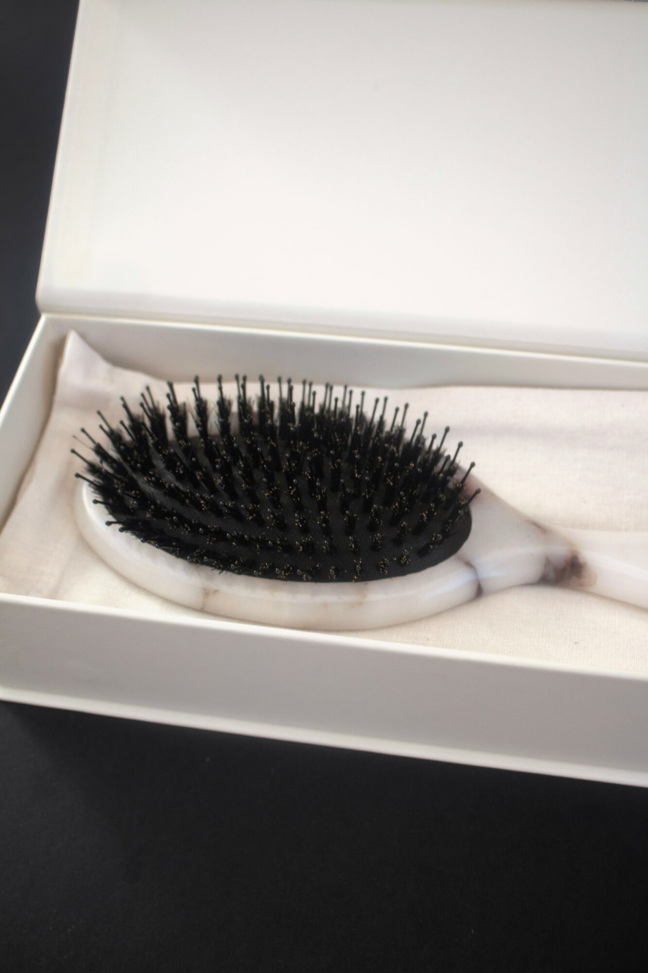 Black hairbrush with a white handle inside a white box, resting on a black surface.