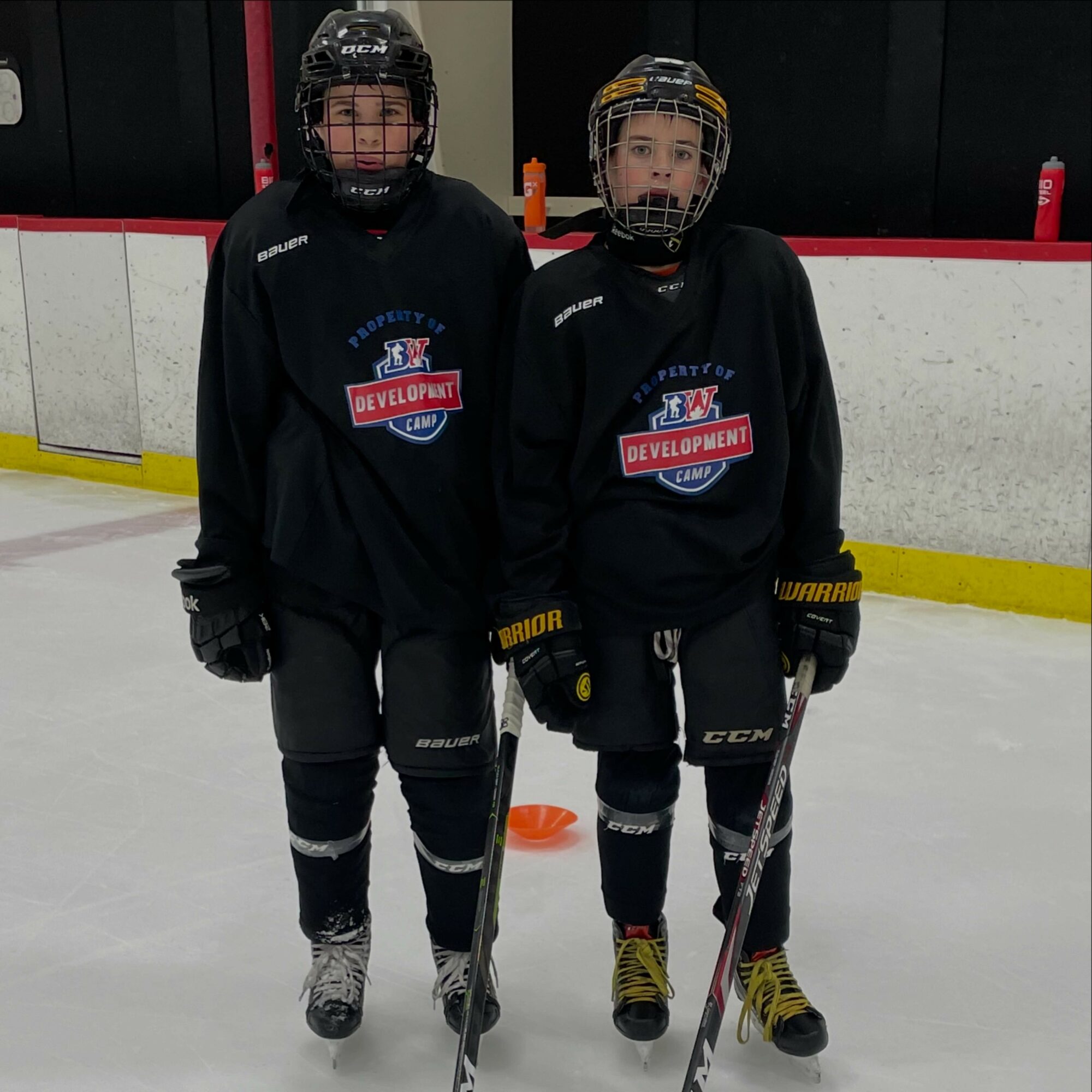 Two young hockey players in black jerseys and helmets stand on ice rink, holding hockey sticks, with a white and red barrier behind them.