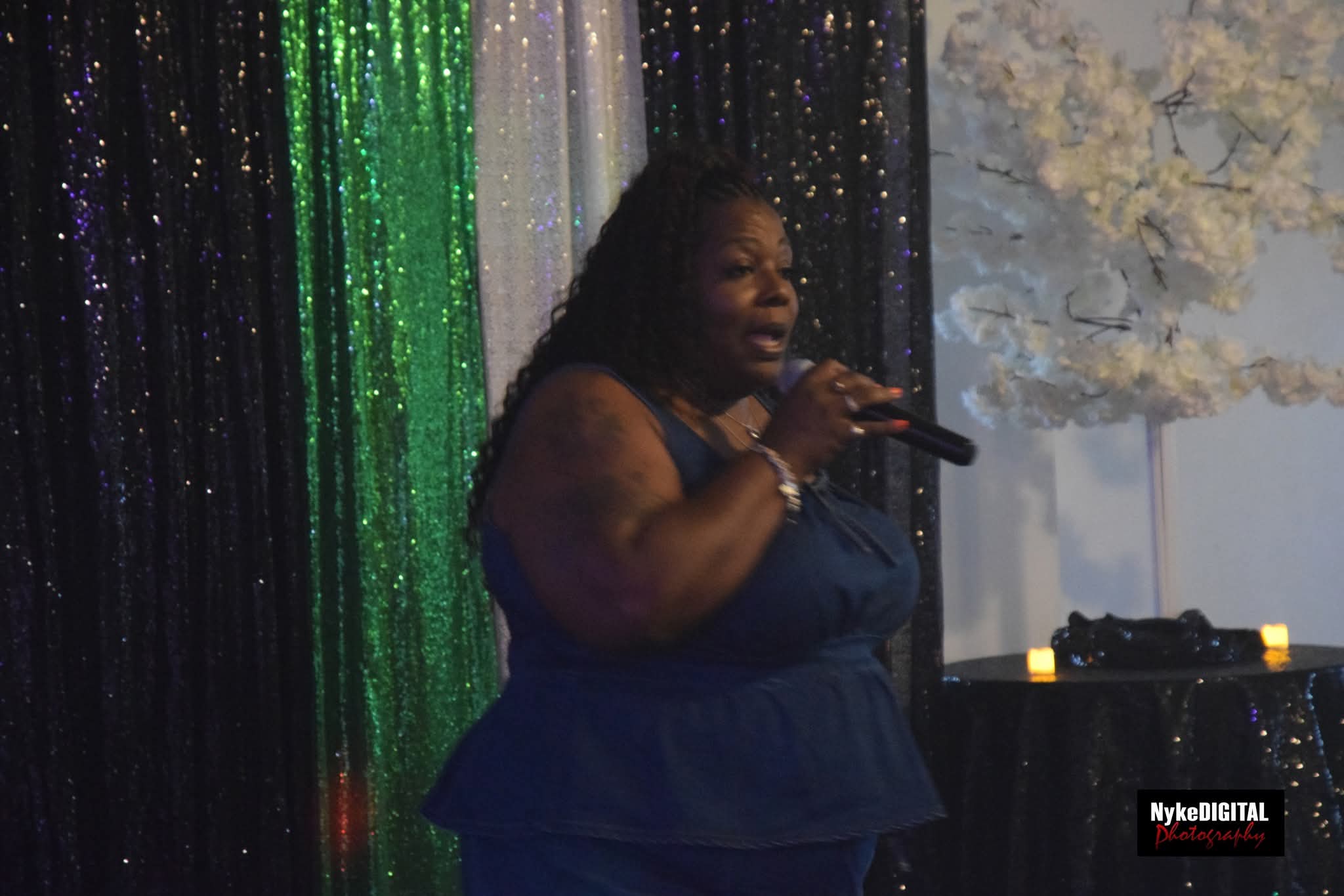 Woman singing into a microphone on stage with colorful backdrop and white floral decoration behind her.