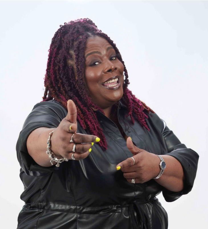 Woman with dreadlocks pointing and smiling, wearing a black jacket, against a plain light background.