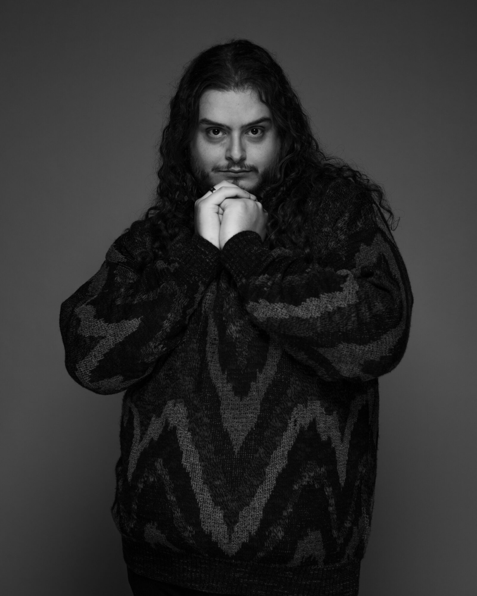 Person with long curly hair and a patterned sweater, hands clasped near face, looking at camera, in black and white.