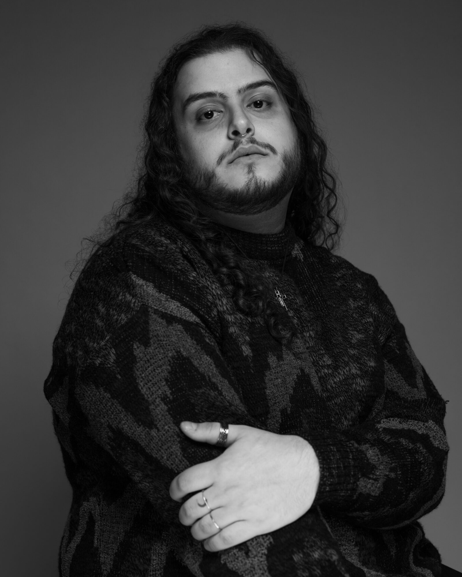 Person with long curly hair and beard, wearing a patterned sweater, looking at the camera with crossed arms.