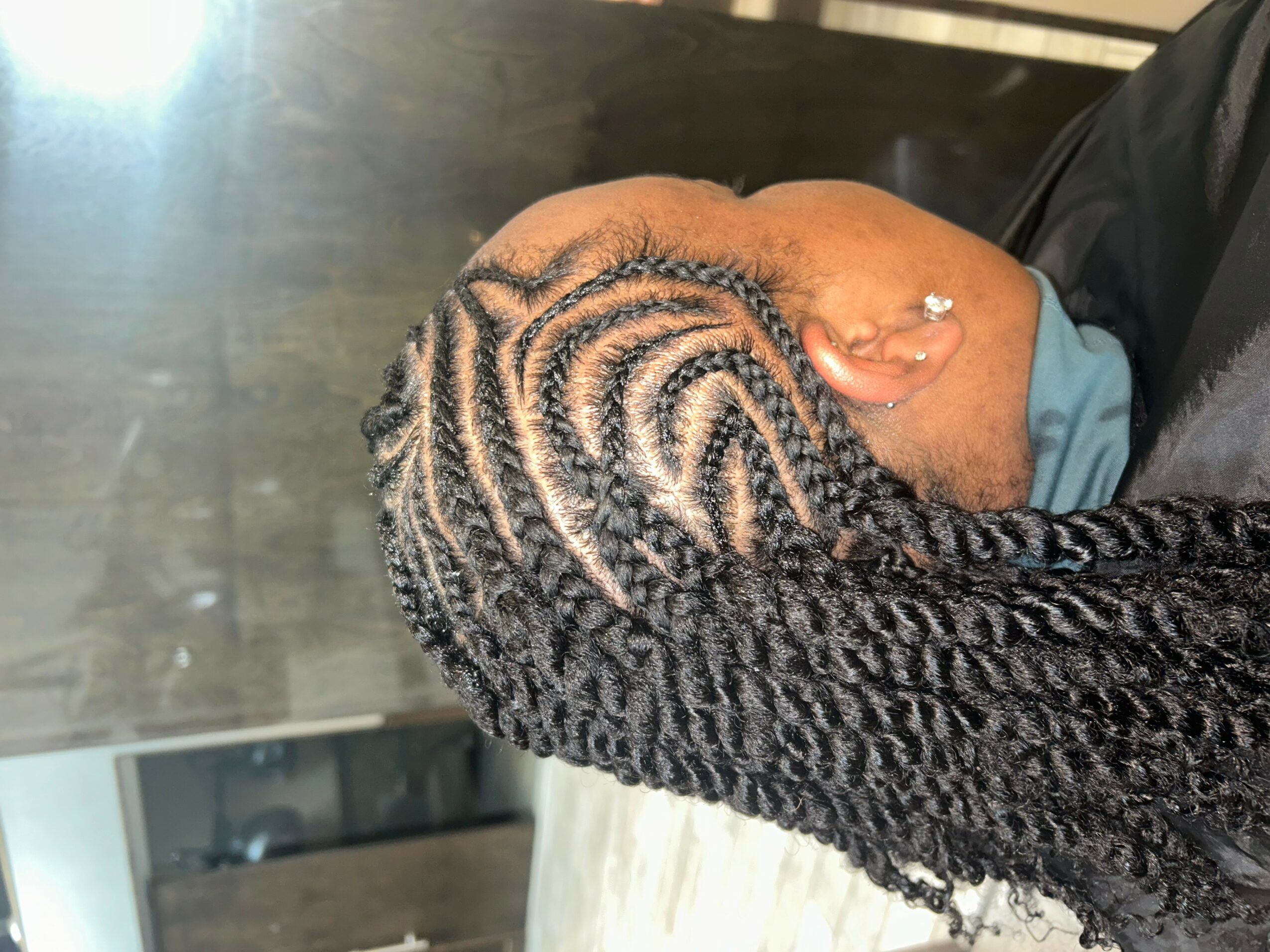 Person with intricately styled braids, head tilted downward, showing detailed hair pattern and a small earring.