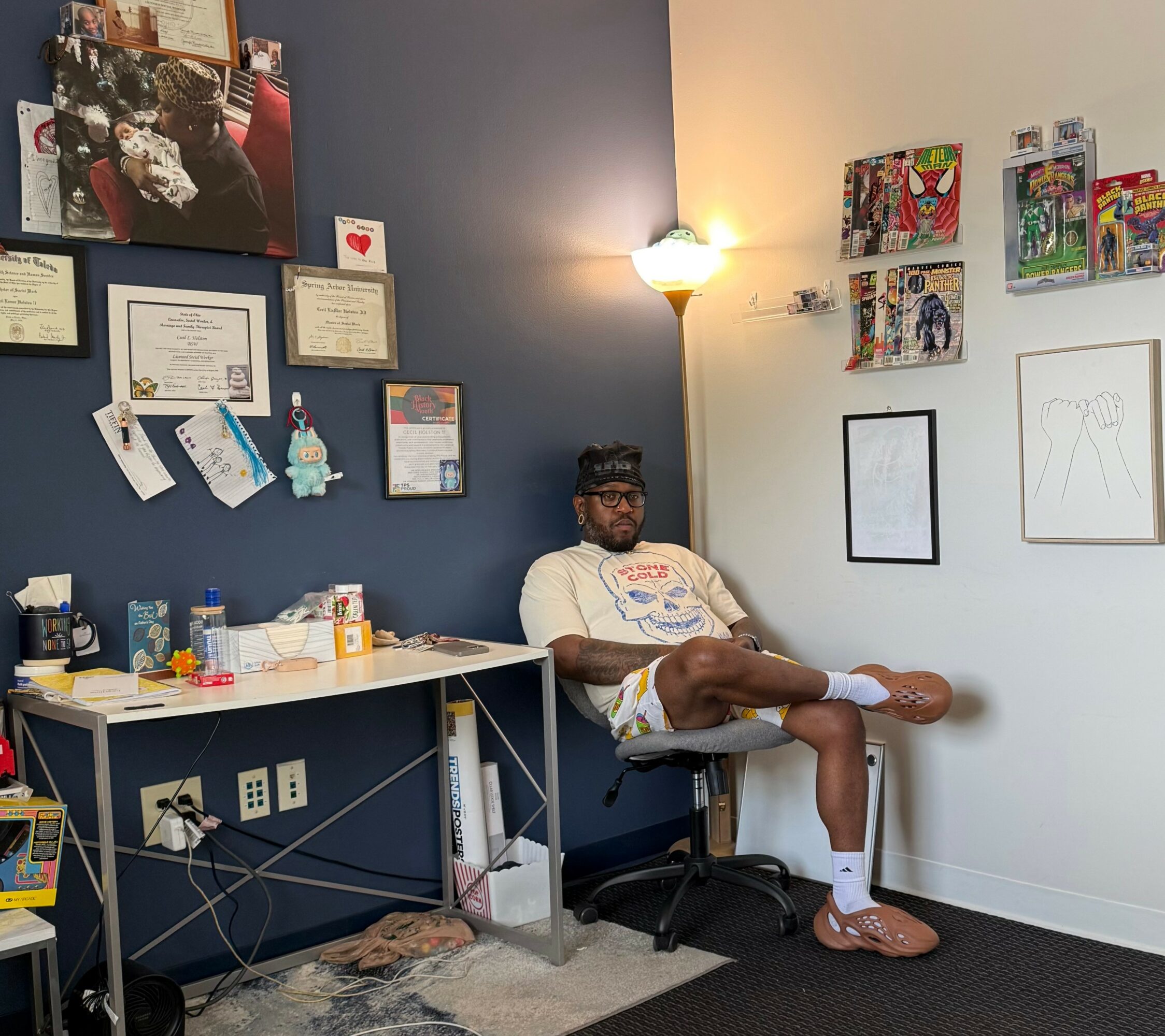 Person sitting in a room with framed certificates and artwork on the walls, a floor lamp, and a desk with various items.