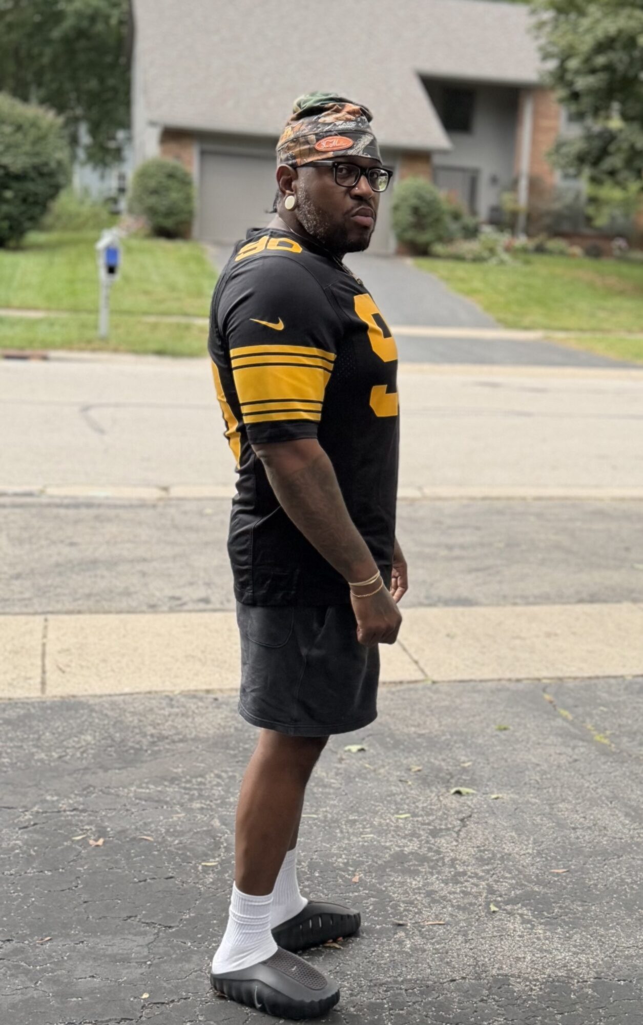 Man standing on sidewalk in black sports jersey, shorts, white socks, and black shoes, with a patterned headscarf, facing sideways.