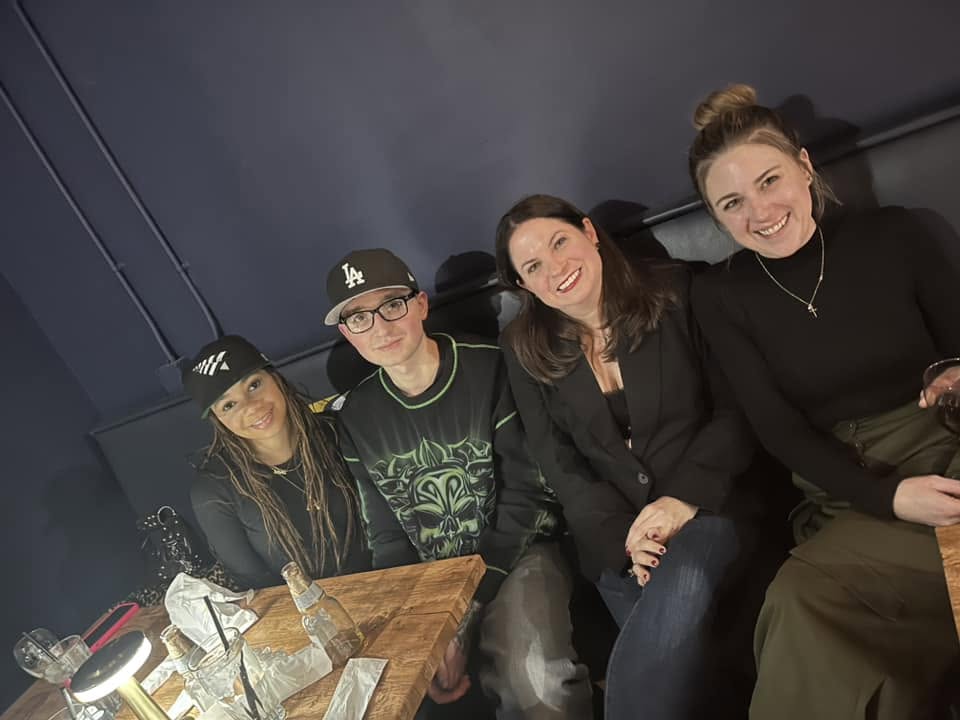 Four people sitting together on a dark-colored couch, smiling, with a wooden table in front of them, in a dimly lit room.