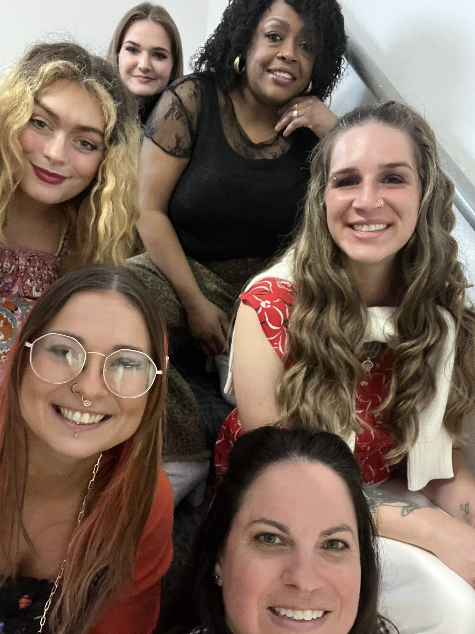 Group of six women smiling, posing closely together indoors, some wearing glasses, diverse hairstyles and clothing styles.