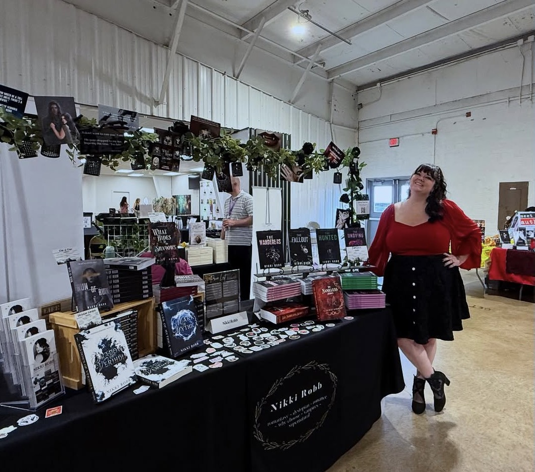 Woman in red top and black skirt stands at a table with books and merchandise in an indoor market. Confident pose.