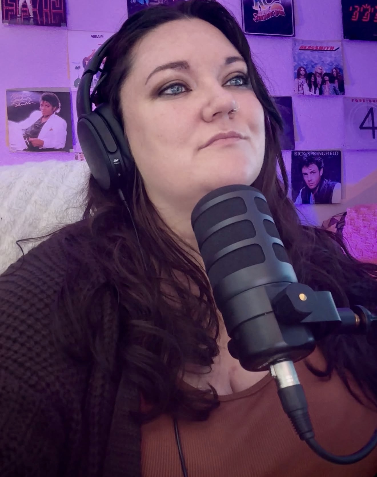 Woman with long dark hair wearing headphones, speaking into a microphone, in a room with posters on the wall.