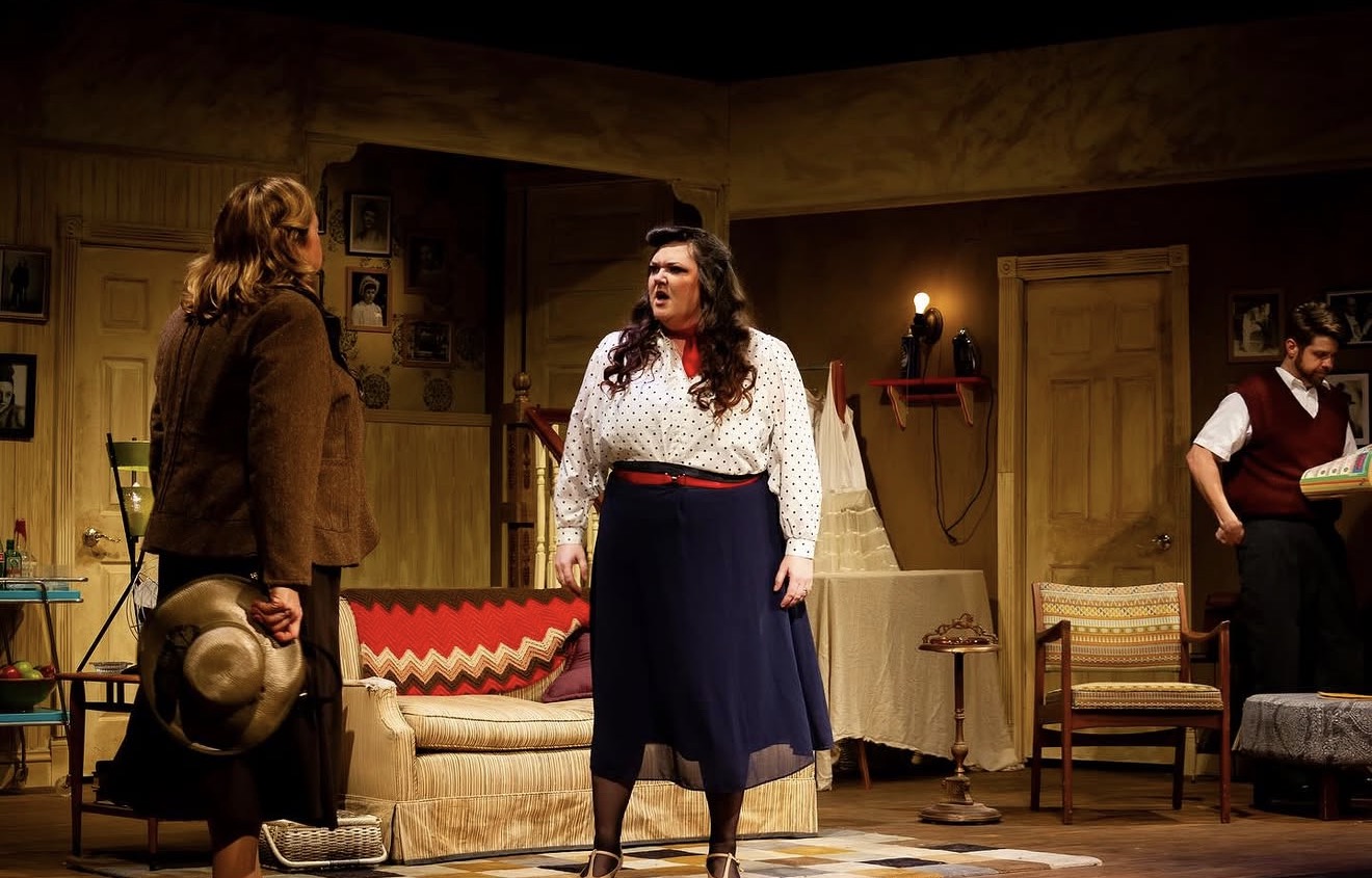 Three people on stage in a living room set, one woman speaking, two others listening, warm lighting, vintage furniture.