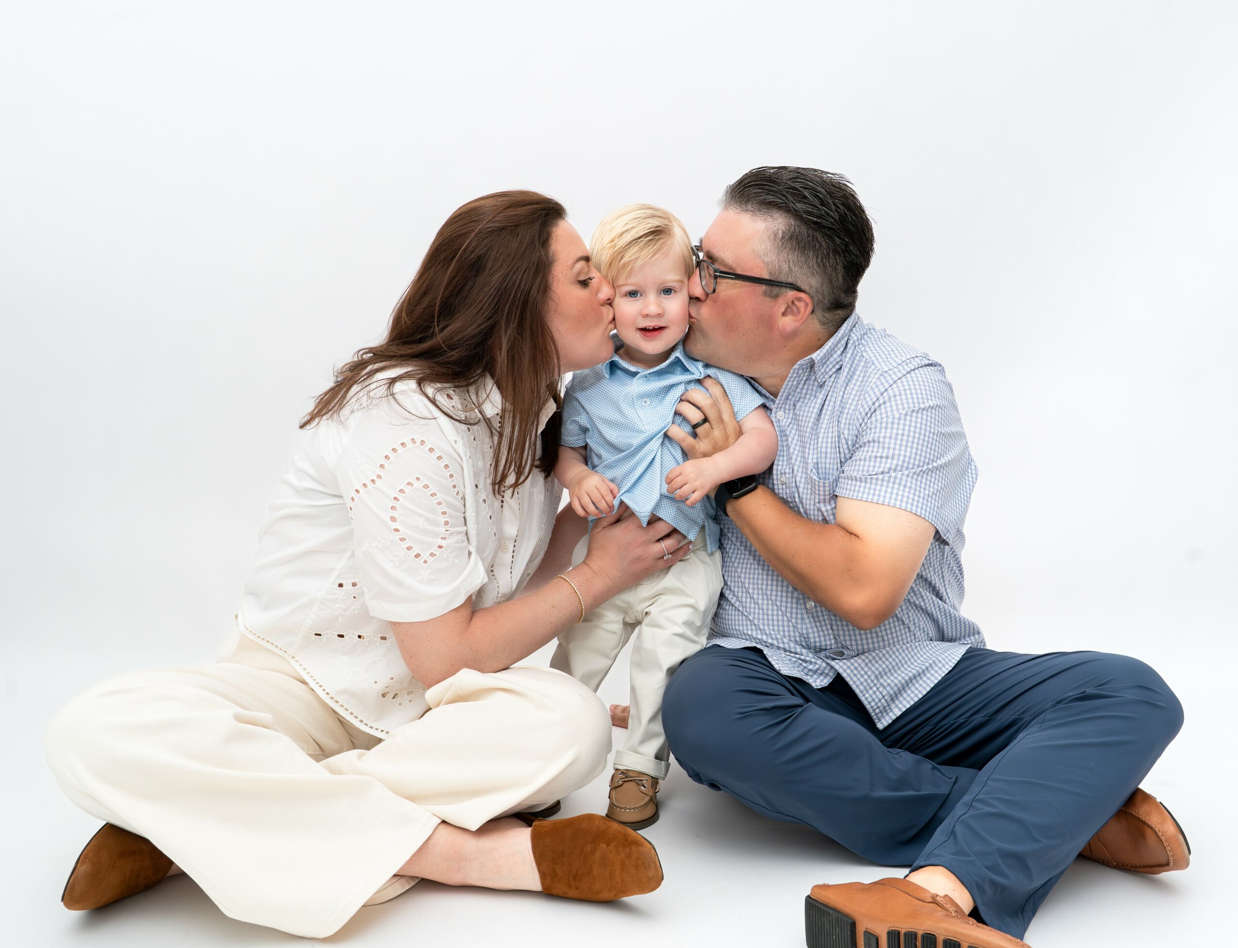 Family of three sitting on the floor, woman and man kissing their child, all smiling, plain white background.