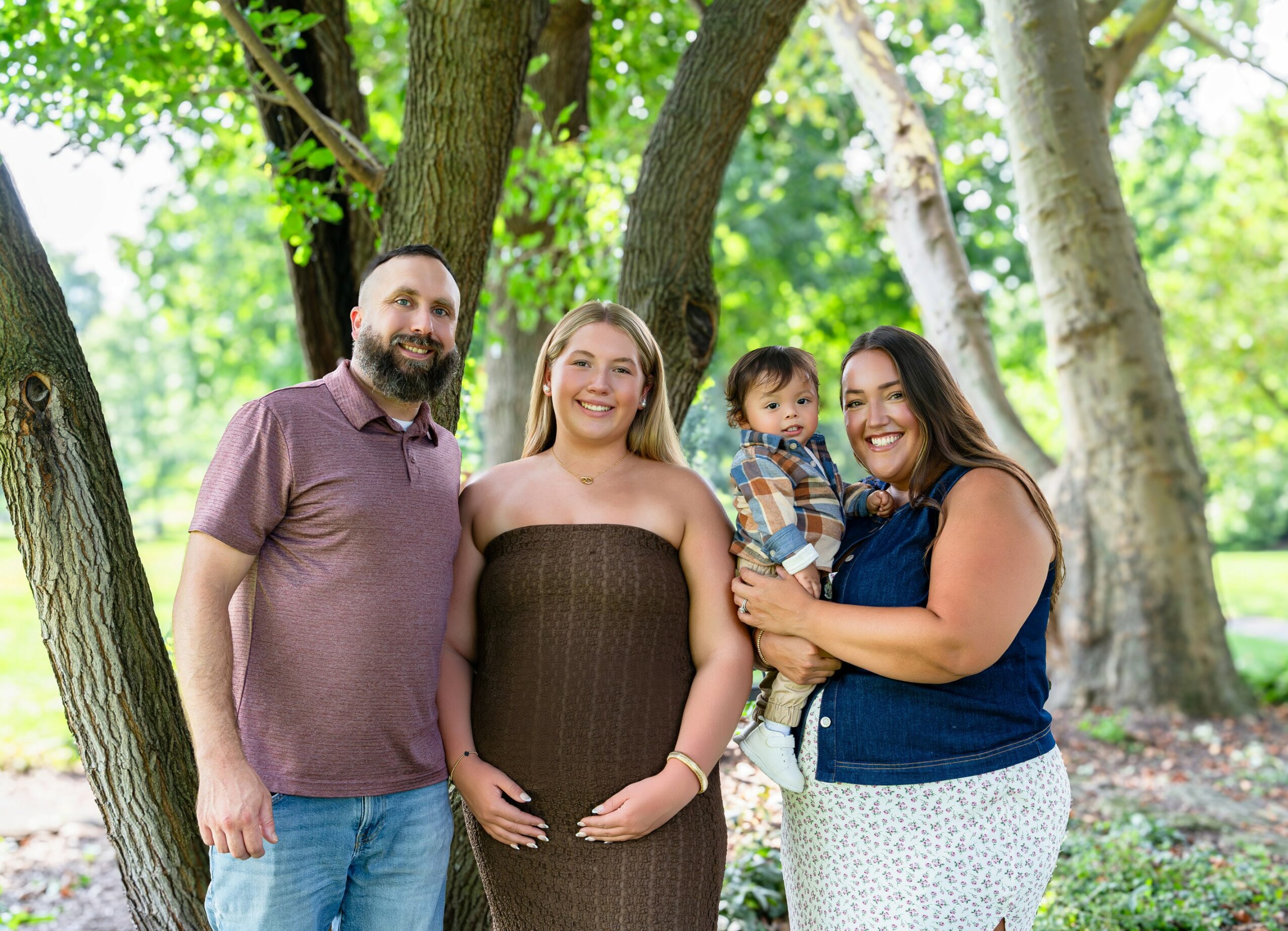Four people standing outdoors among trees, smiling, with a woman holding a child, in a park setting.