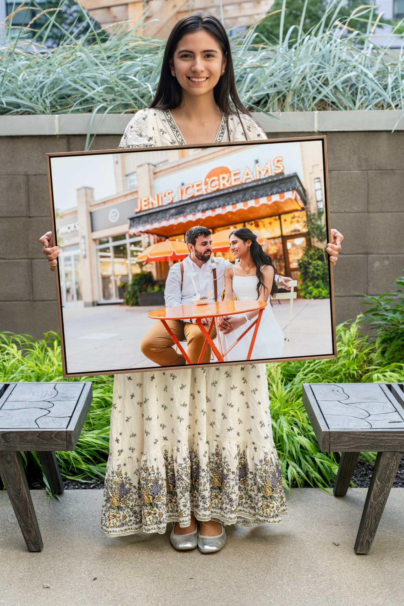 Young woman in a floral dress holding a large framed photo outdoors, with plants and benches in the background.