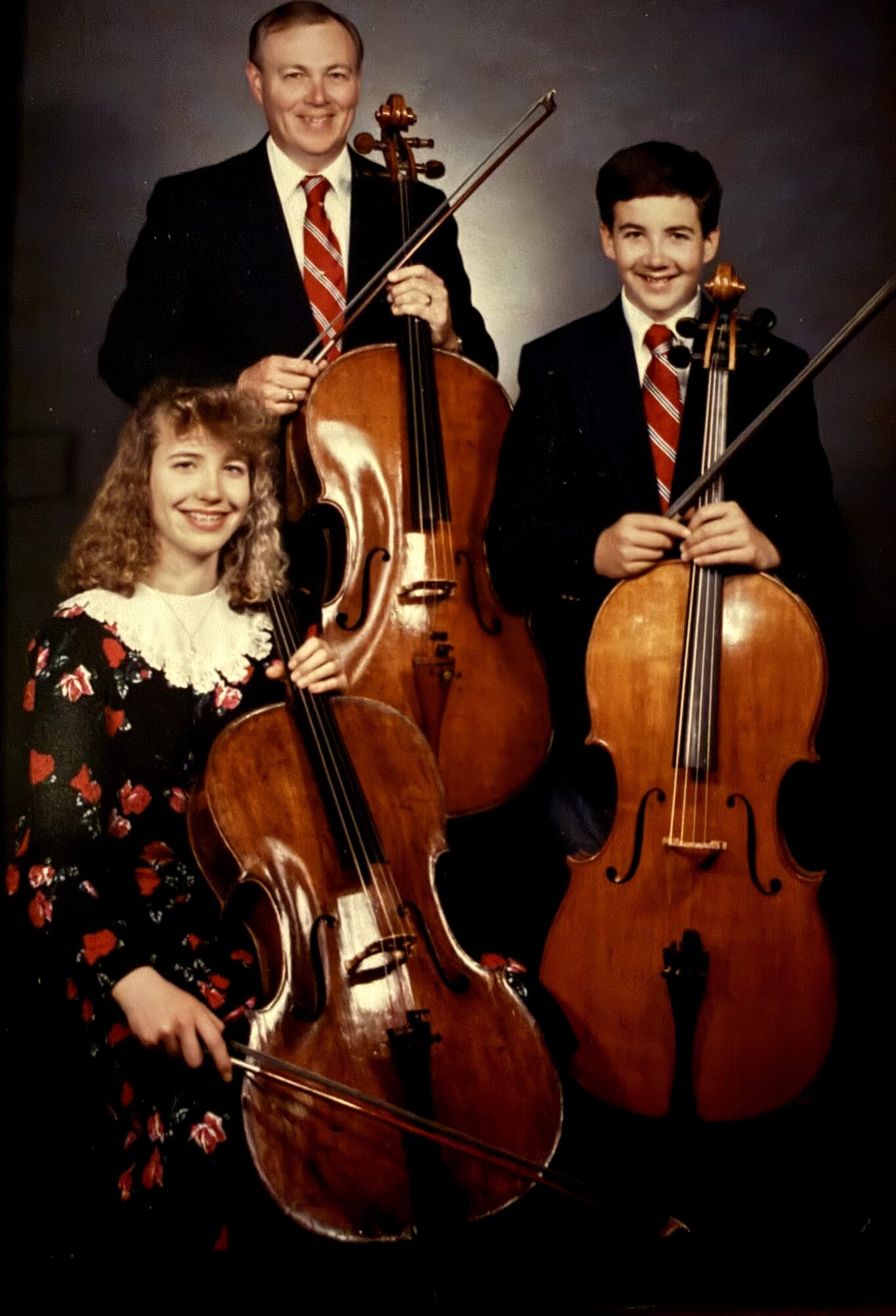 Three people pose with a cello and two violins against a dark studio backdrop.