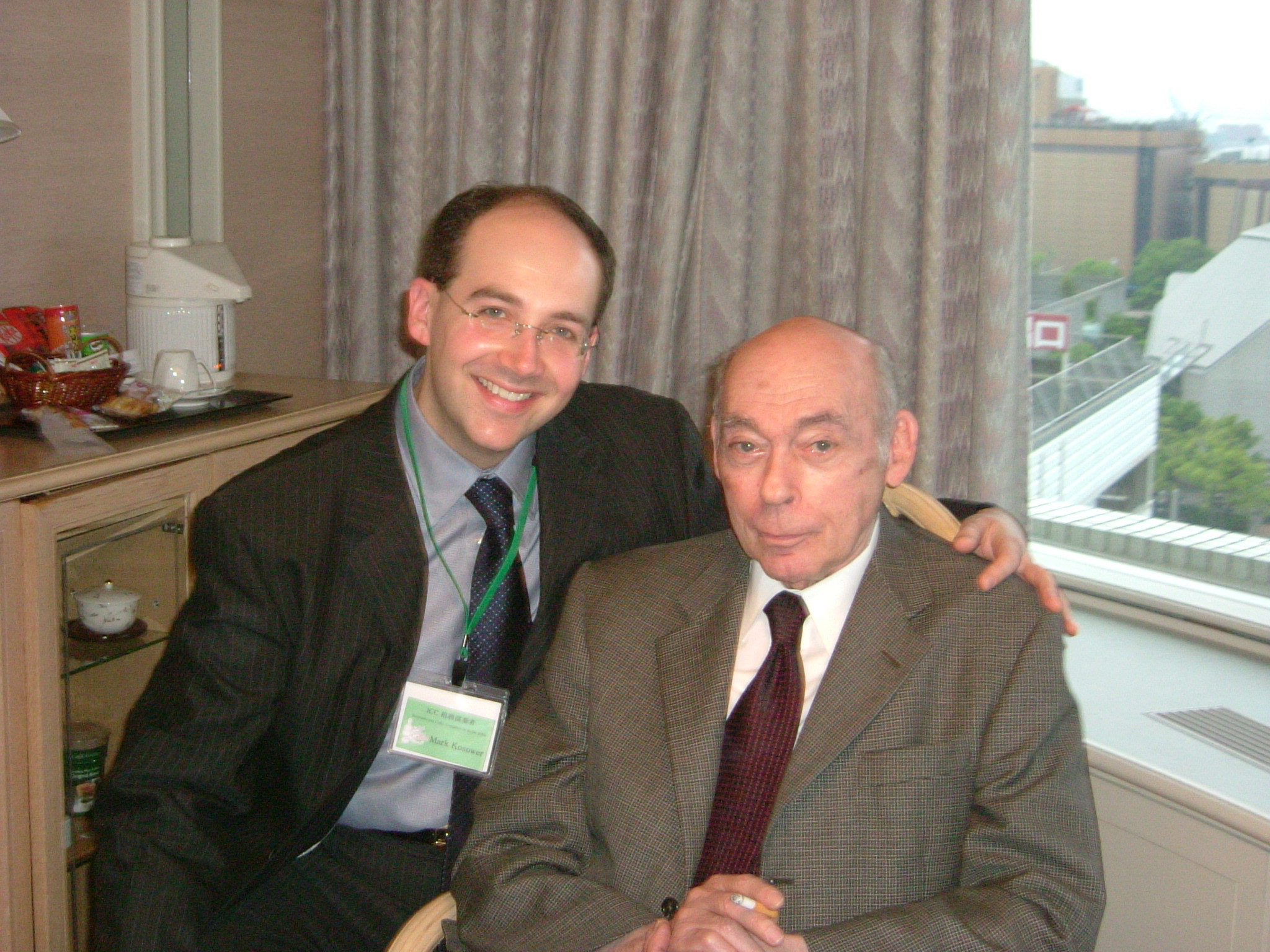 Two men in suits sit together indoors beside a window and curtains.