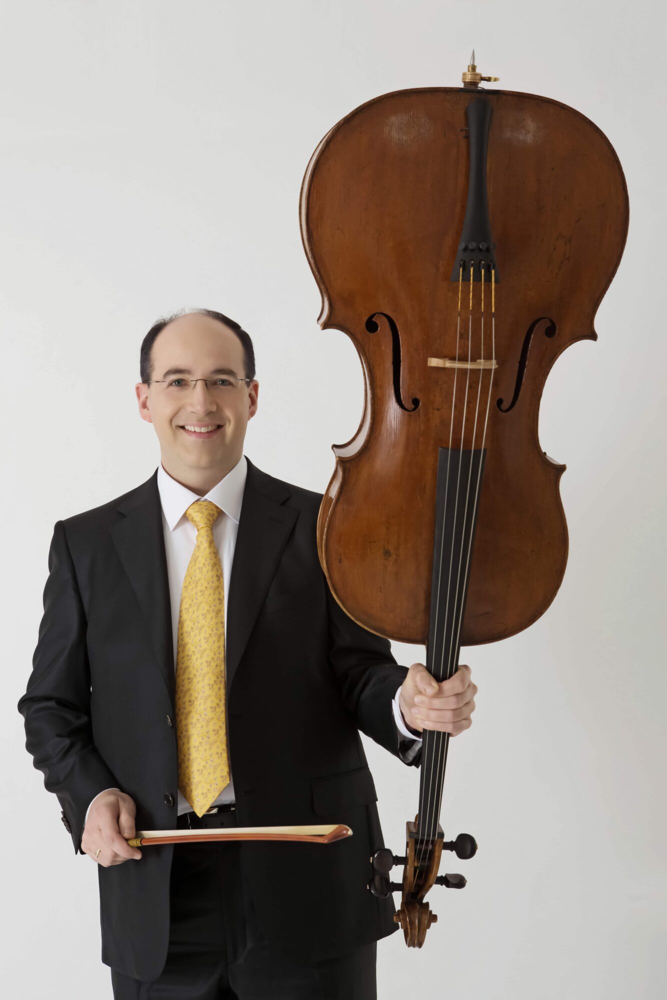 A smiling person in a dark suit holds a cello and bow against a plain background.