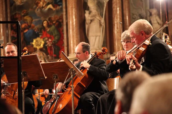 Orchestra musicians play string instruments in an ornate hall, with seated audience members in the foreground.