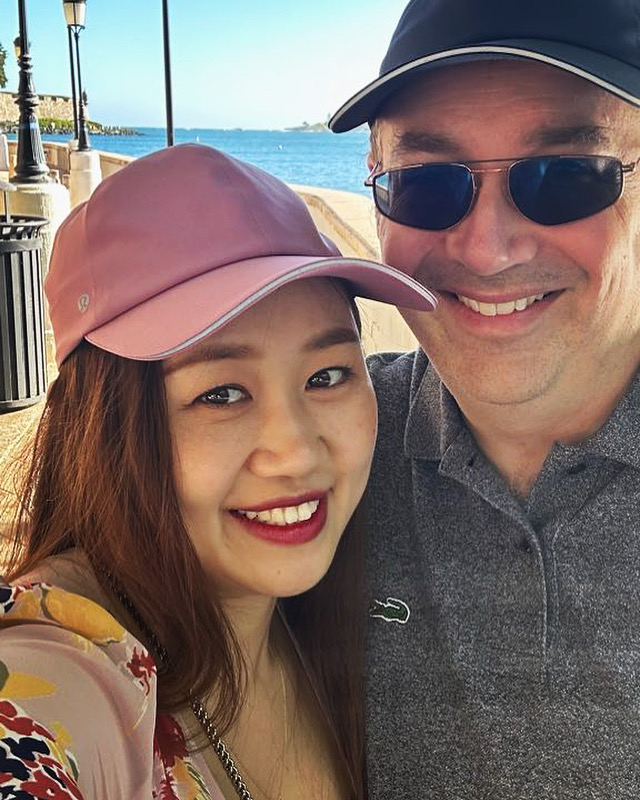 Two people wearing caps and sunglasses pose together beside a waterfront.