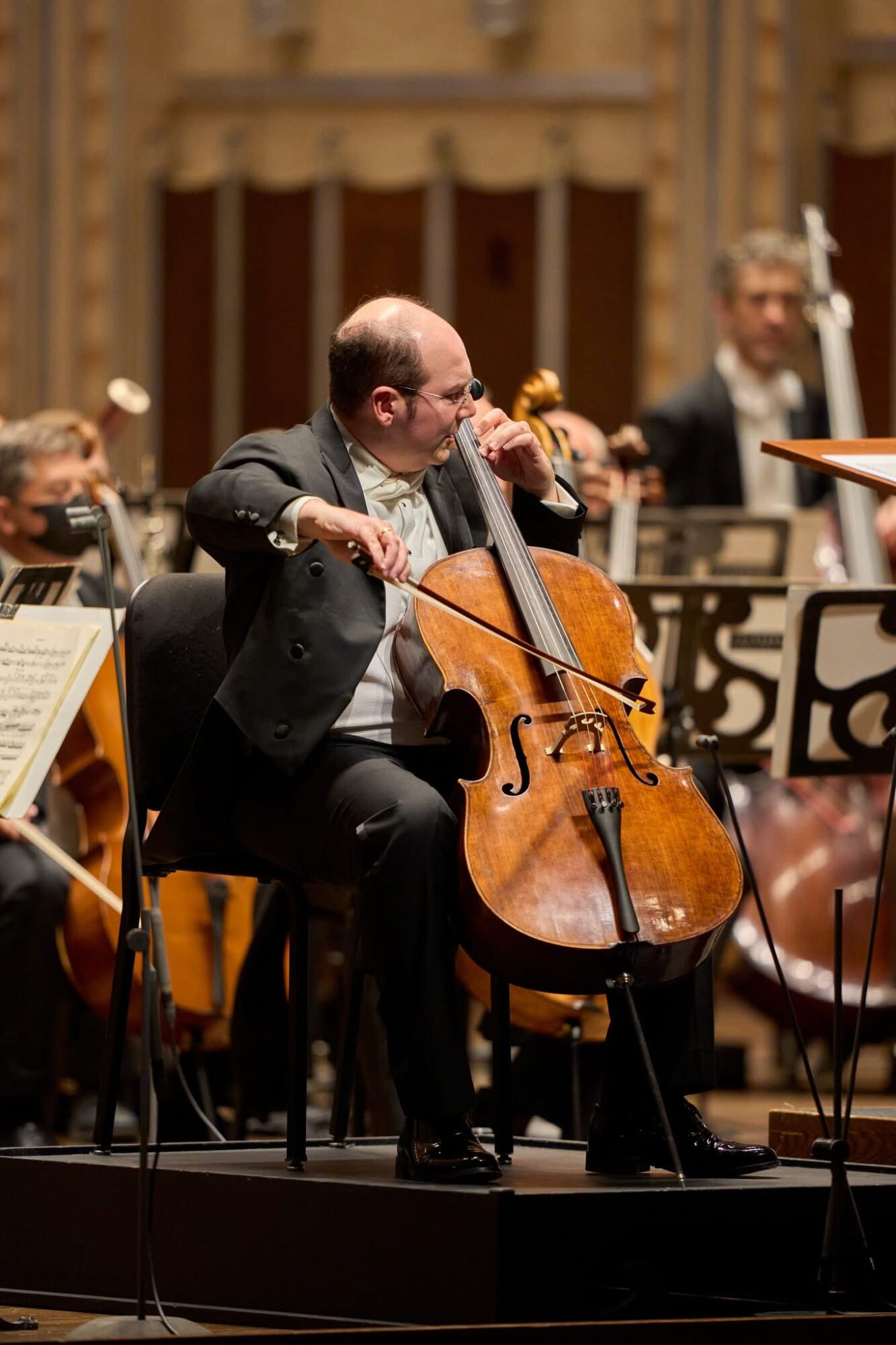 A seated cellist plays a cello onstage while musicians and music stands are visible behind him.