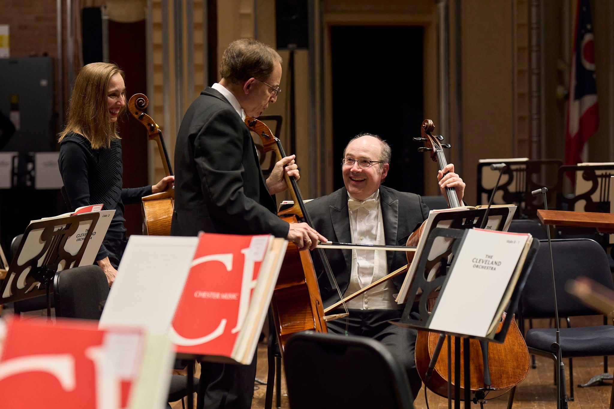 Three musicians perform with string instruments on a stage, with sheet music in the foreground.