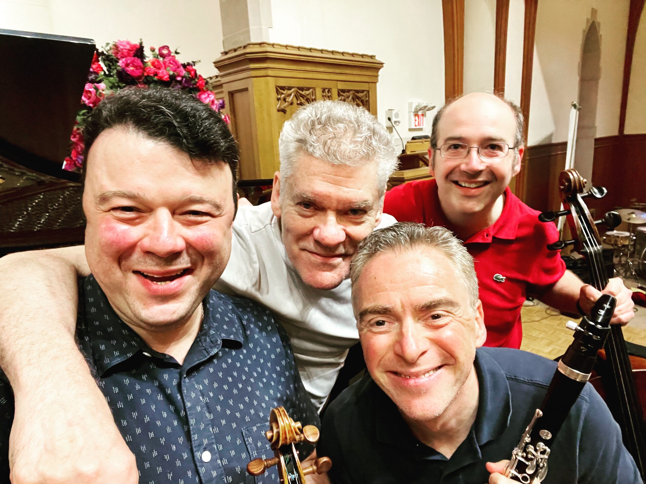 Four men pose together in a music room, with violins and other instruments nearby.
