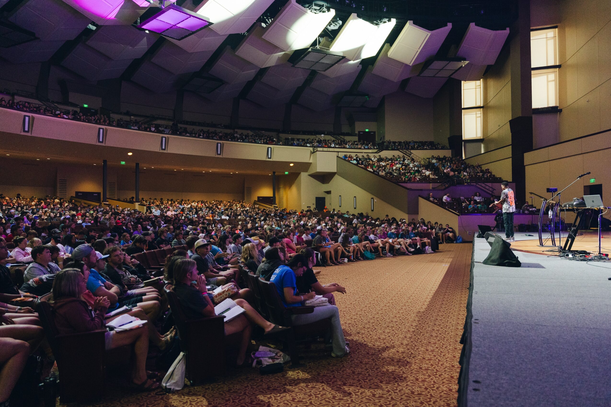 A large audience seated in an auditorium faces a stage with speakers and musical equipment.