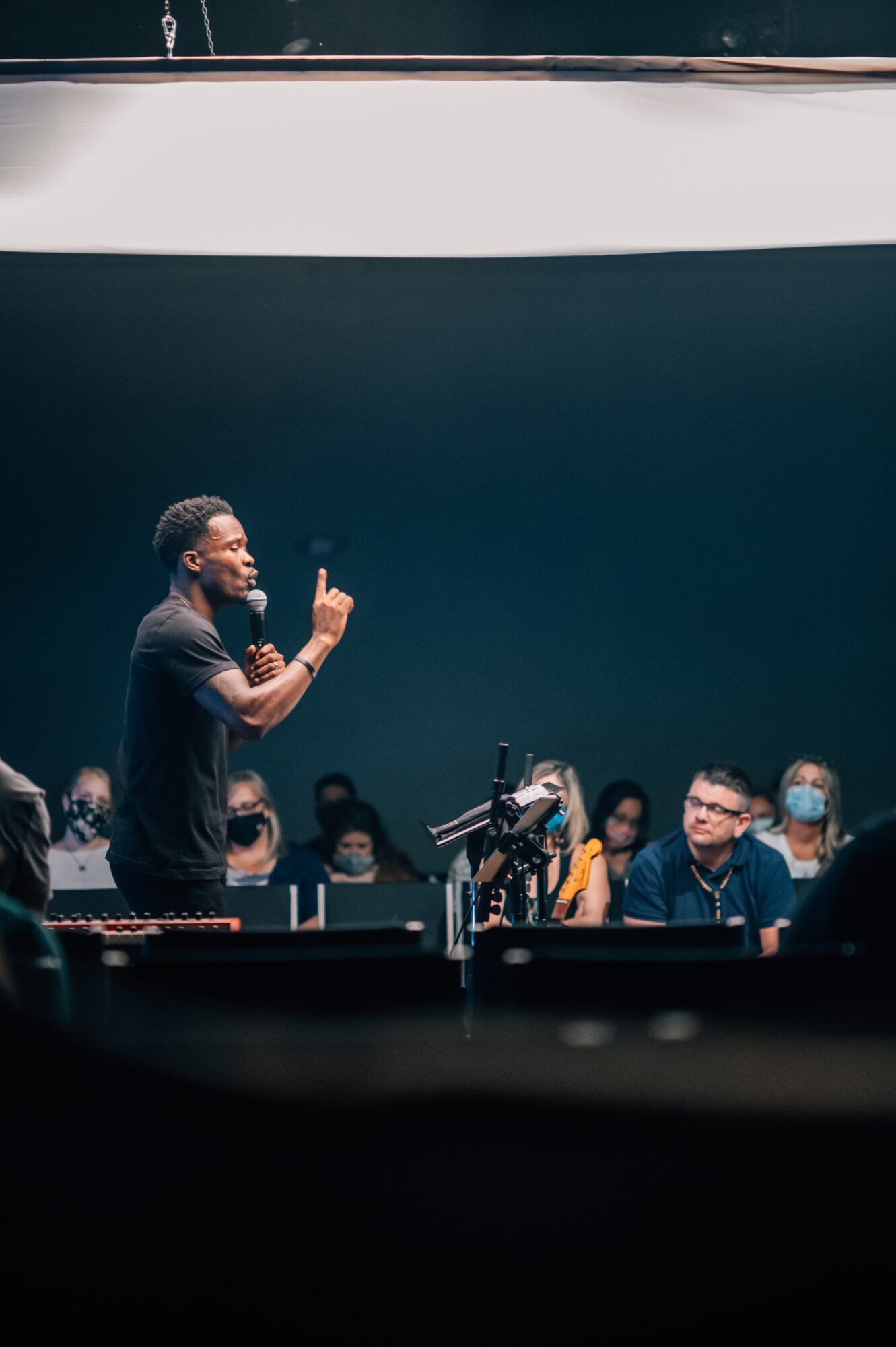 A speaker gestures toward an audience seated in a dim auditorium.