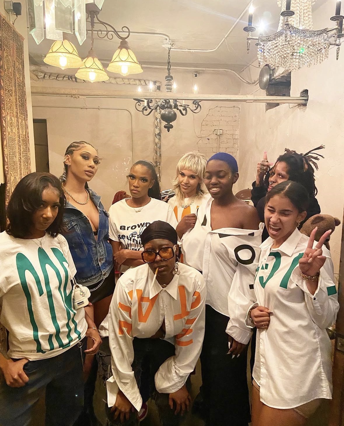 Group of eight women posing indoors, some making peace signs, in a room with chandeliers and warm lighting.