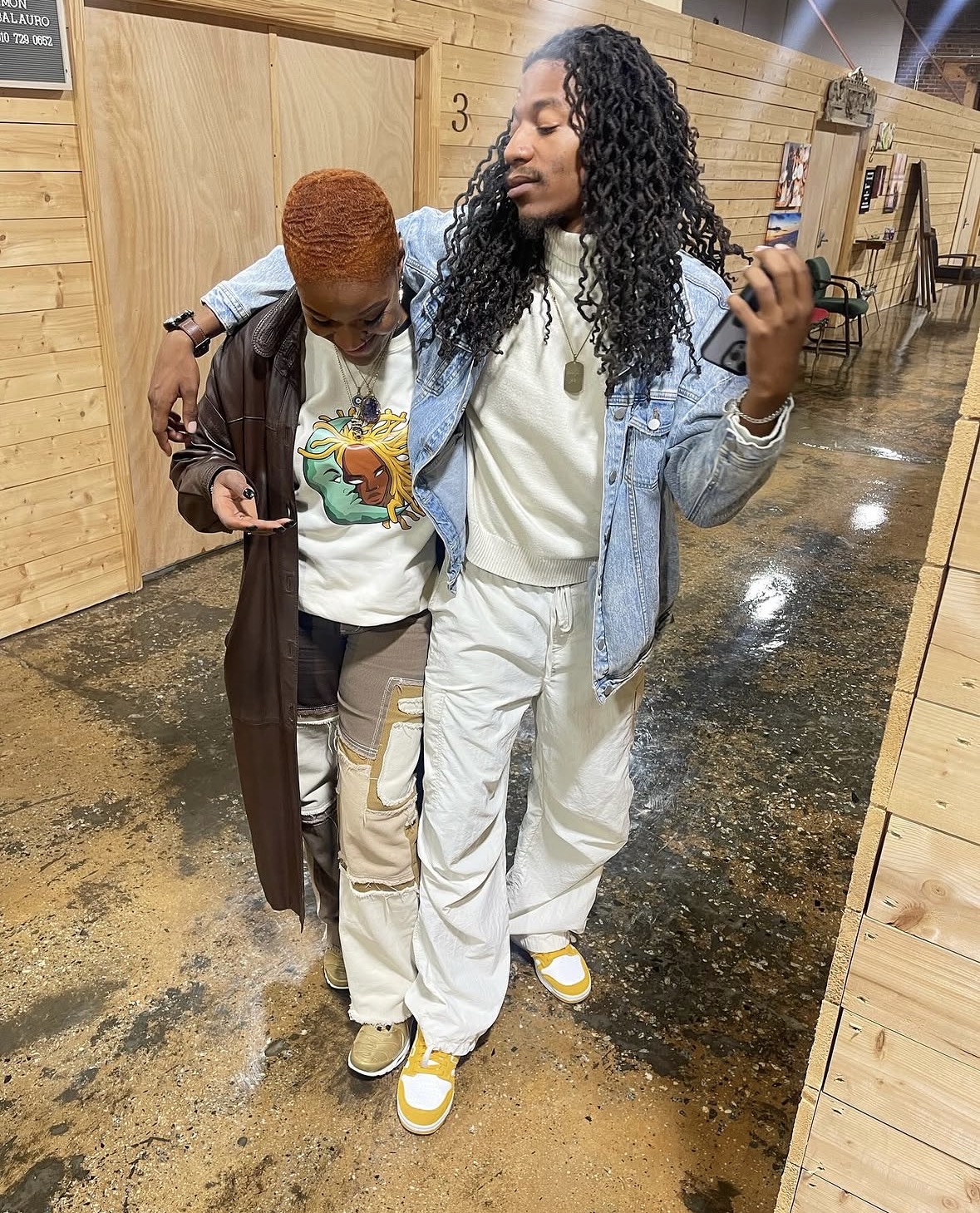 Two women walk together in a wooden-floored indoor space, one with curly hair and the other with short hair, both dressed casually.