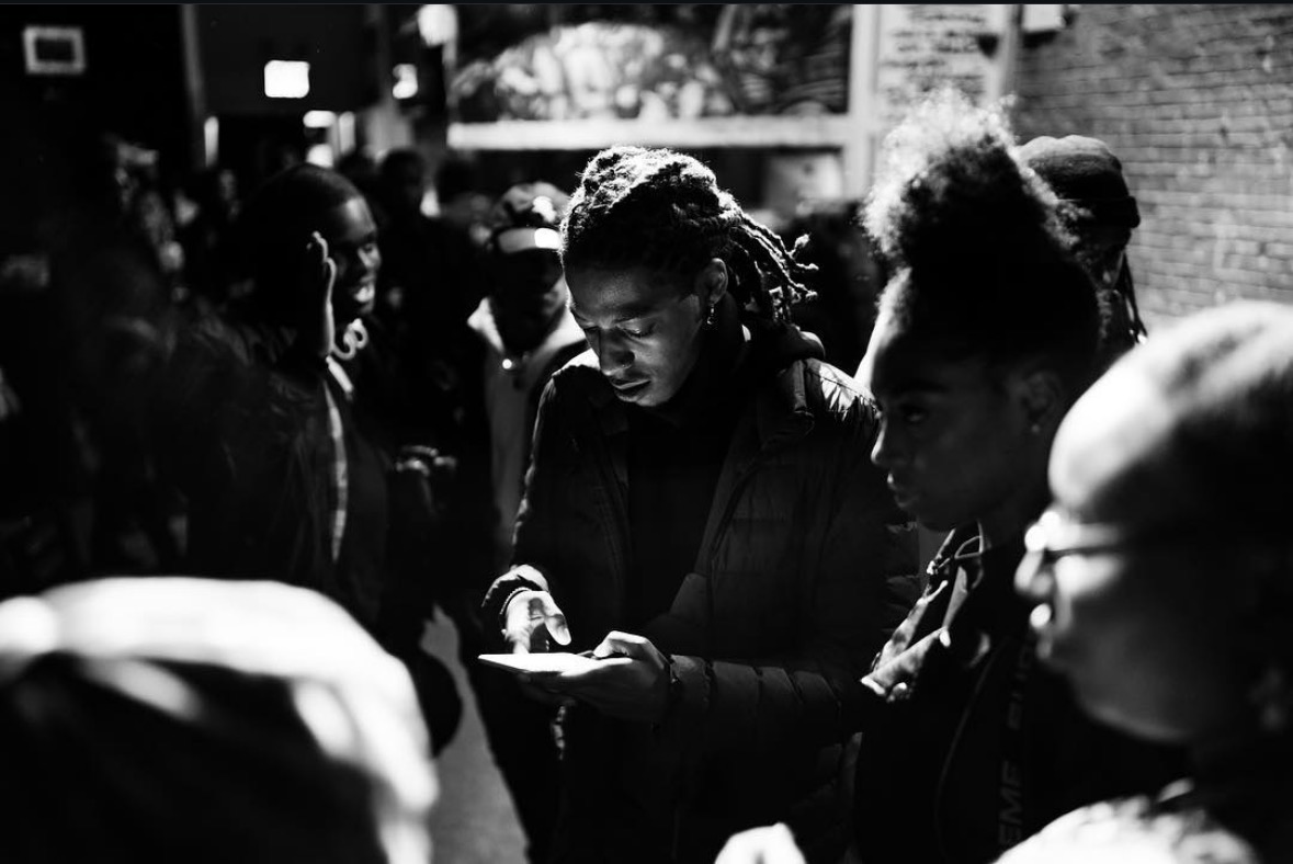 Group of people gathered outdoors, some looking at a phone, in black and white.