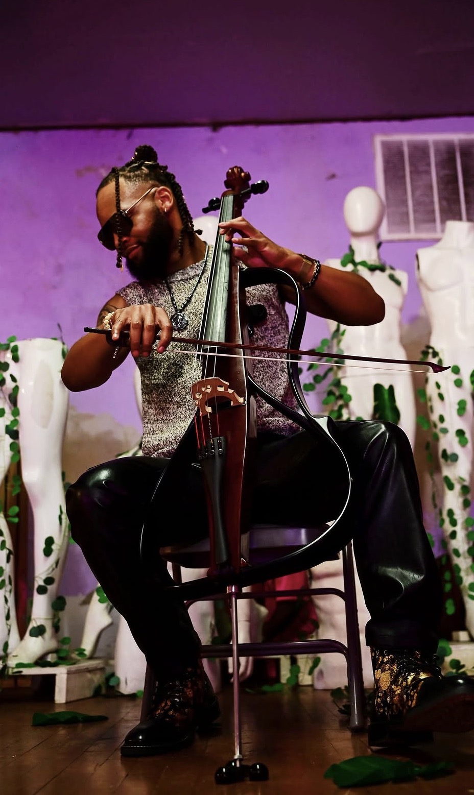 Person playing a cello on stage with purple background and white decorative elements, wearing glasses and a patterned shirt.