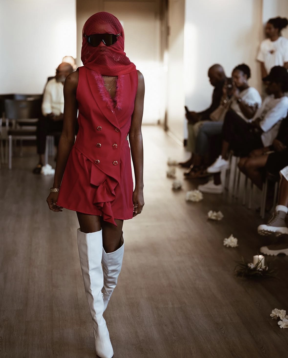 Person walking down runway wearing red dress, white thigh-high boots, sunglasses, and red head covering, audience seated on sides.