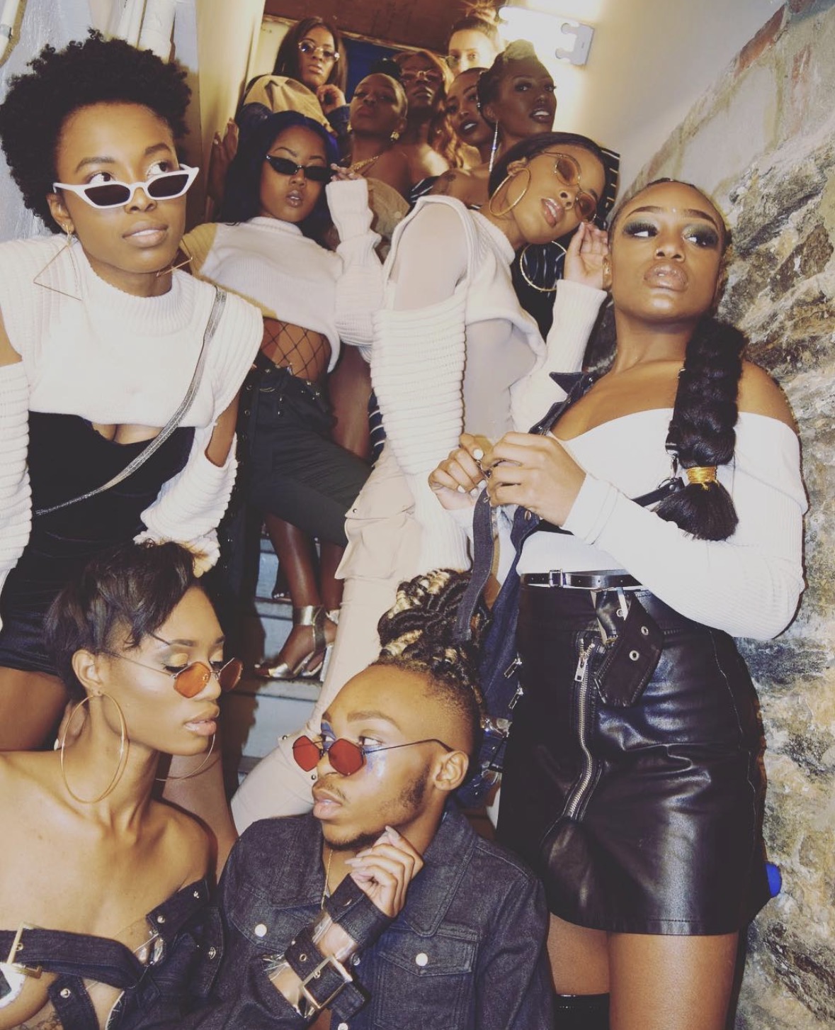 Group of women posing indoors, some wearing sunglasses, stylish outfits, and accessories, with a stone wall in the background.