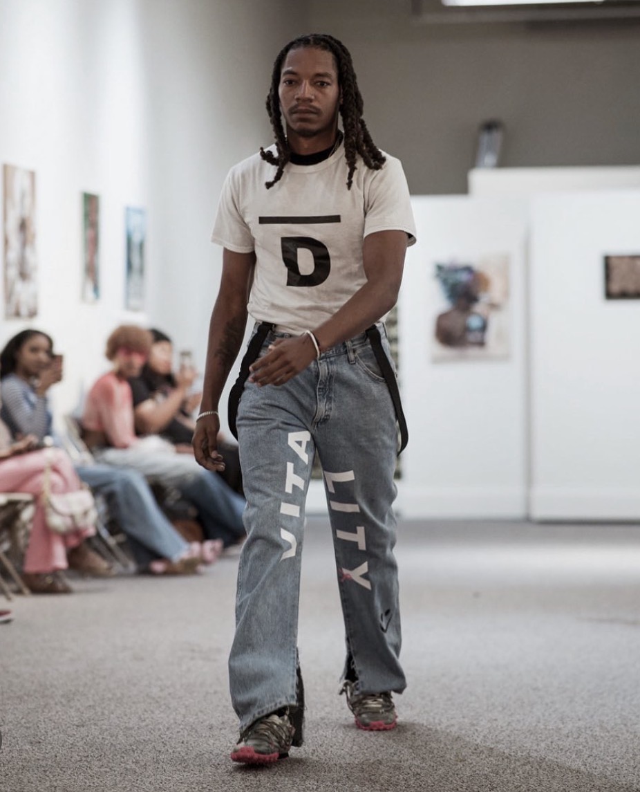 Young man with dreadlocks walking in an art gallery, with people seated and artwork displayed on walls behind him.