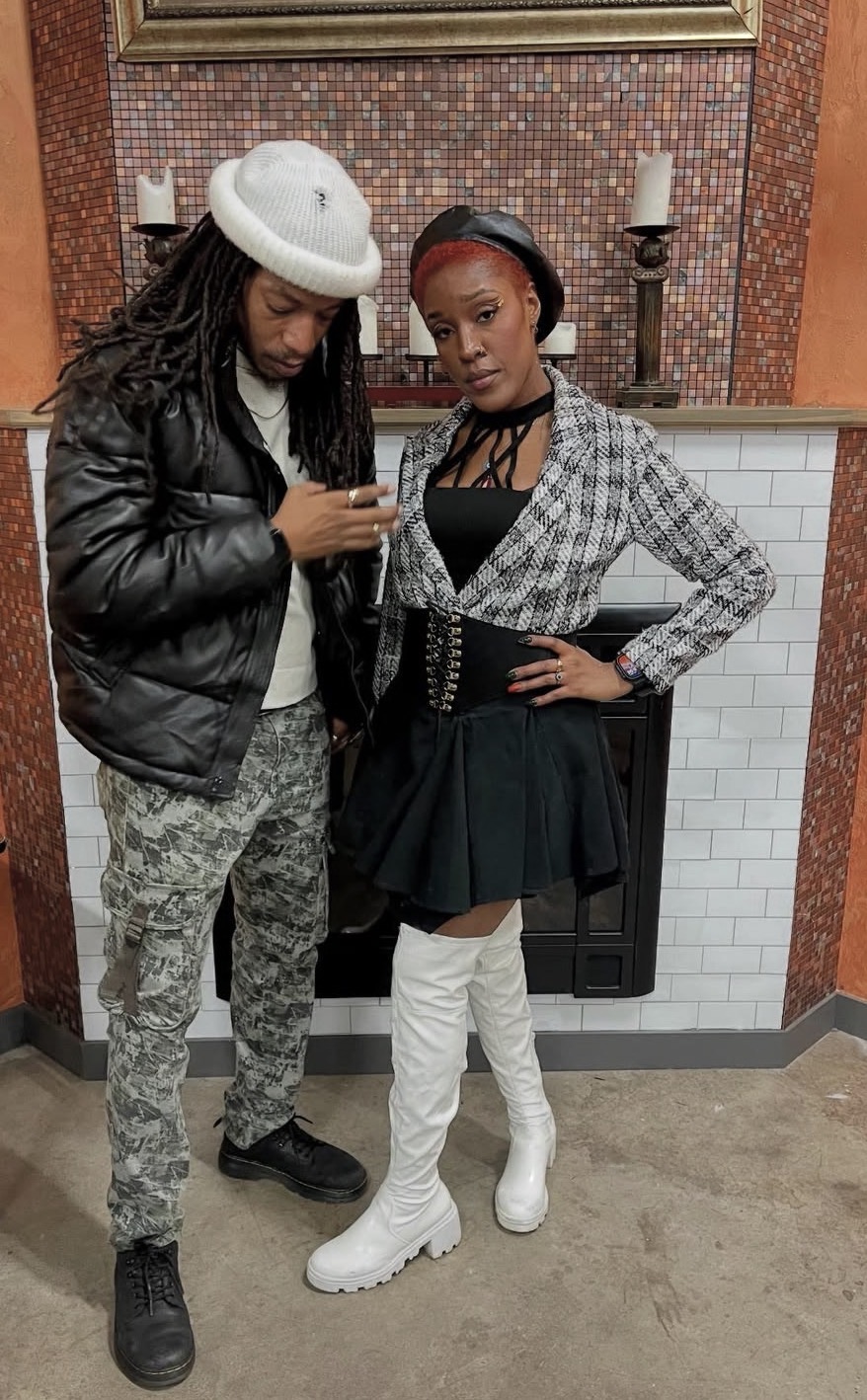 Two women standing in front of a fireplace, one looking at her phone, the other posing with hand on hip, both dressed fashionably.