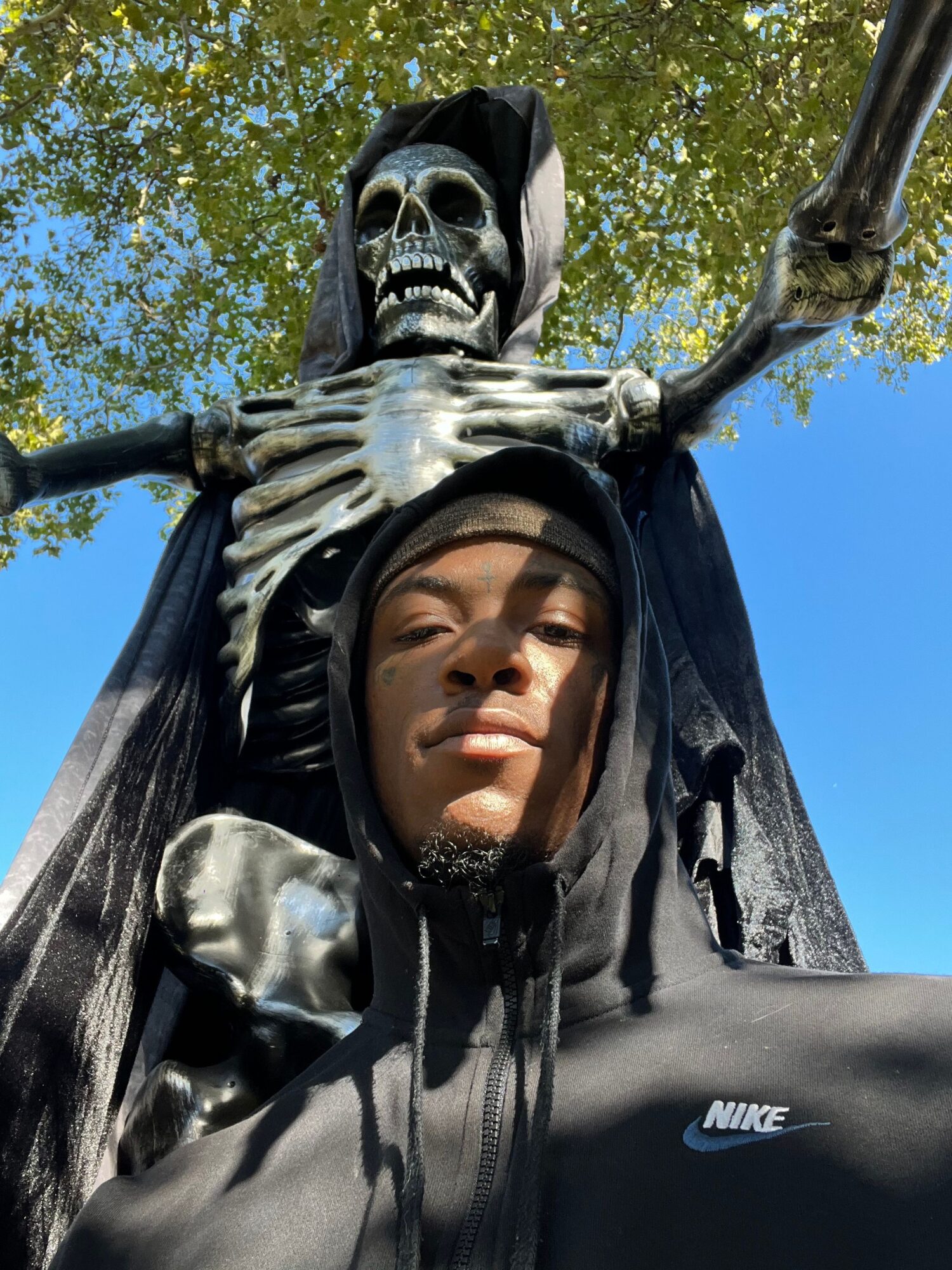 Person in a black Nike jacket poses beside a skeletal statue outdoors beneath a tree.