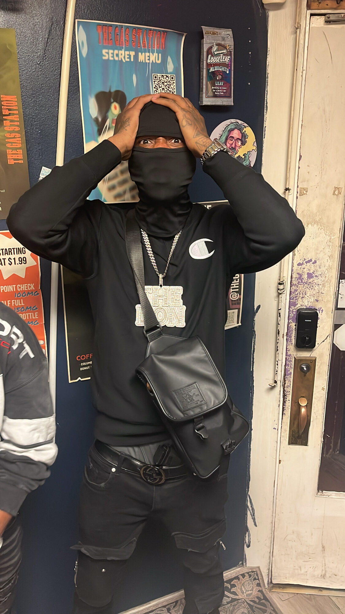 Person wearing dark clothing and a face covering stands with hands on their head in front of posters and a door.