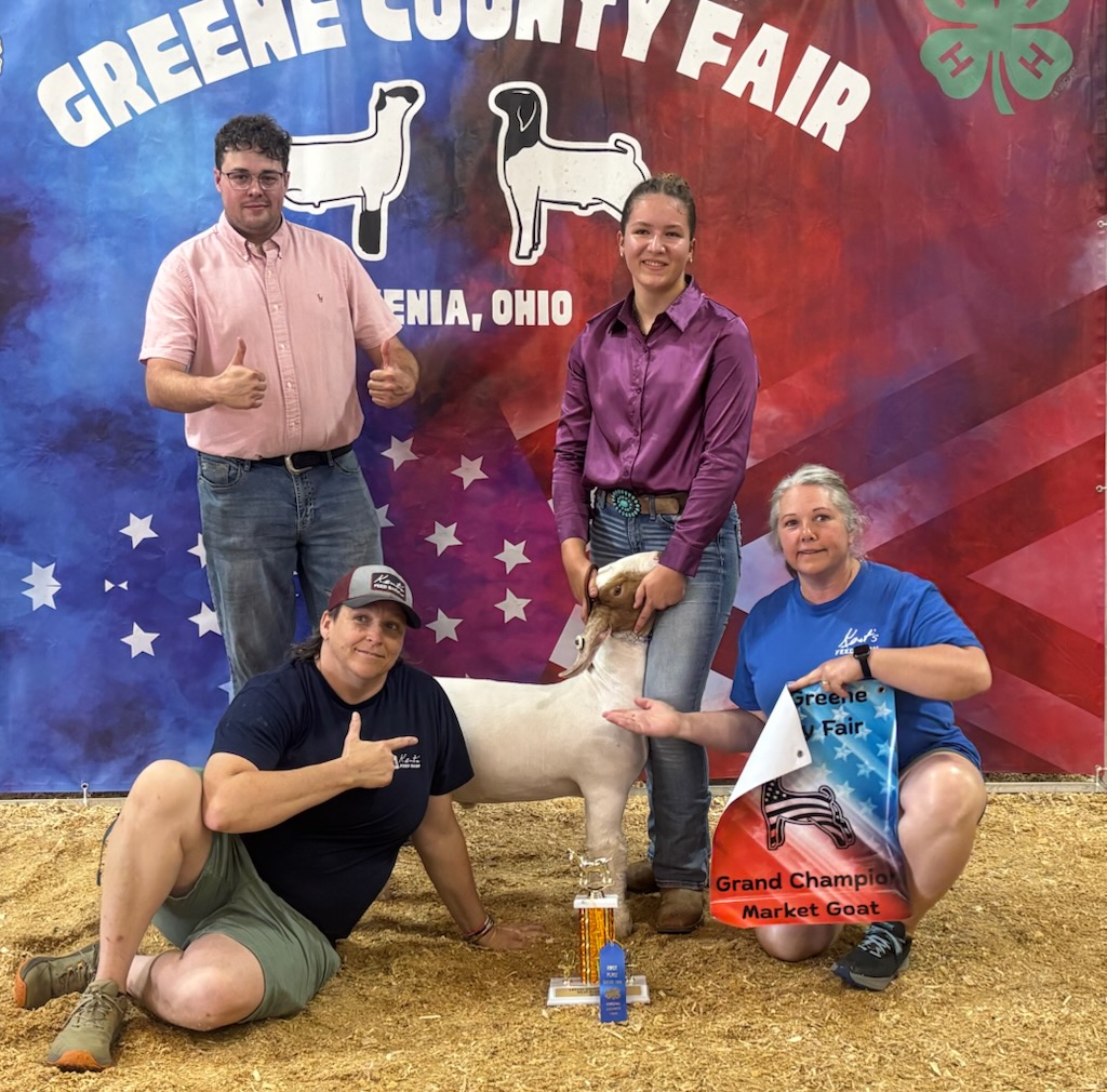 Four people pose with a goat at Greene County Fair, with a banner in the background. One person holds a trophy.