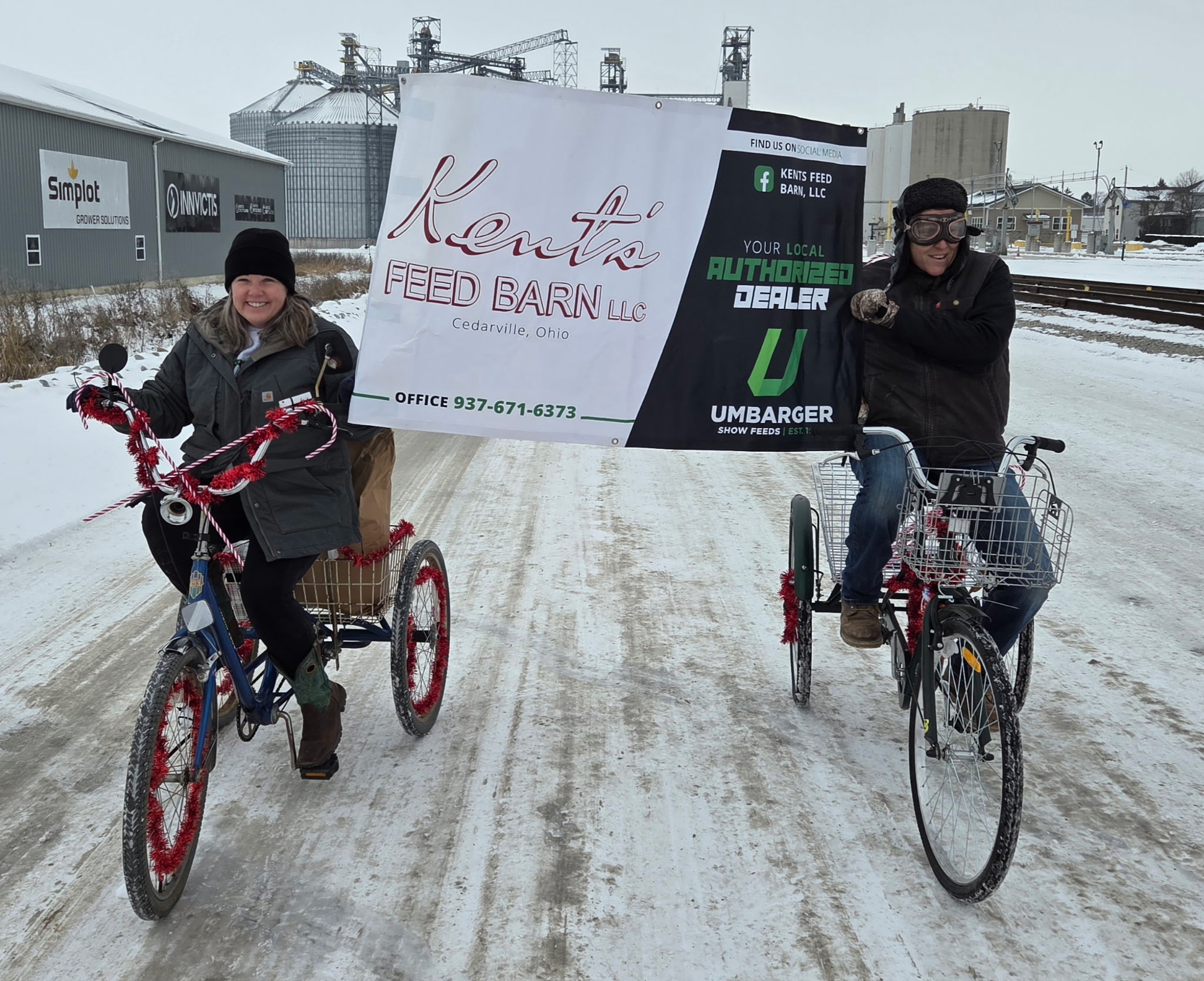 Two people on bicycles holding a large banner outdoors in snowy weather, with industrial buildings in the background.