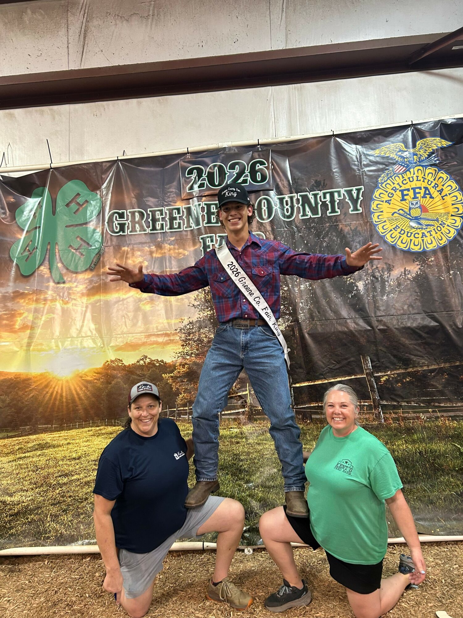 Three people standing outdoors in front of a banner, with one person in the middle holding a trophy, all smiling.
