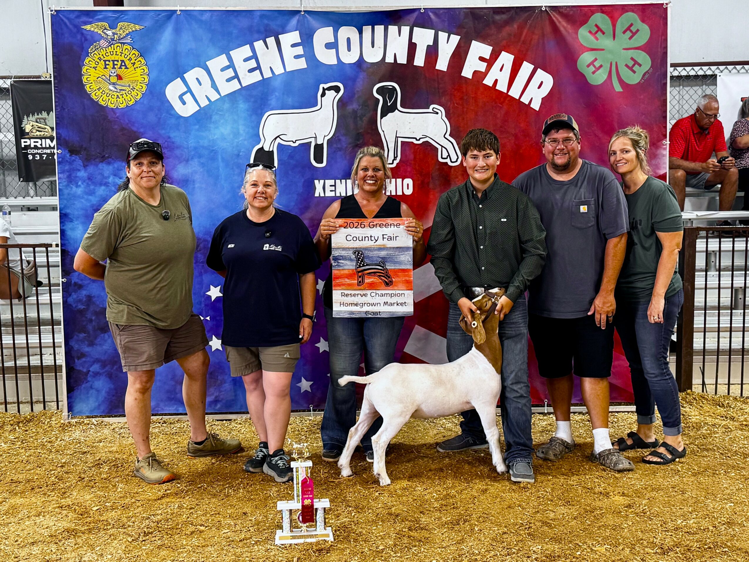 Group of six people standing with a white goat at Greene County Fair, holding a ribbon and prize. Banner in background.