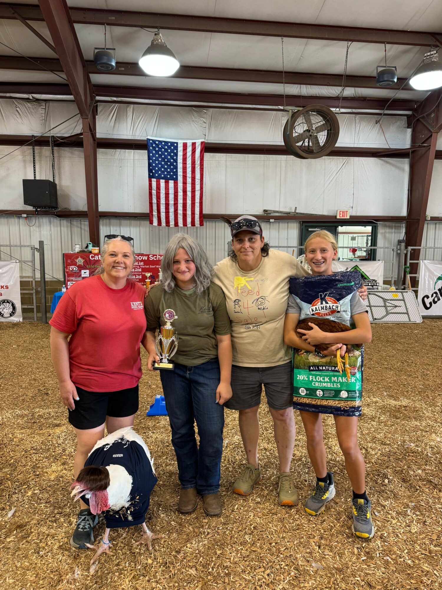 Four women and a girl standing indoors with an American flag, some holding trophies, in a barn-like setting.