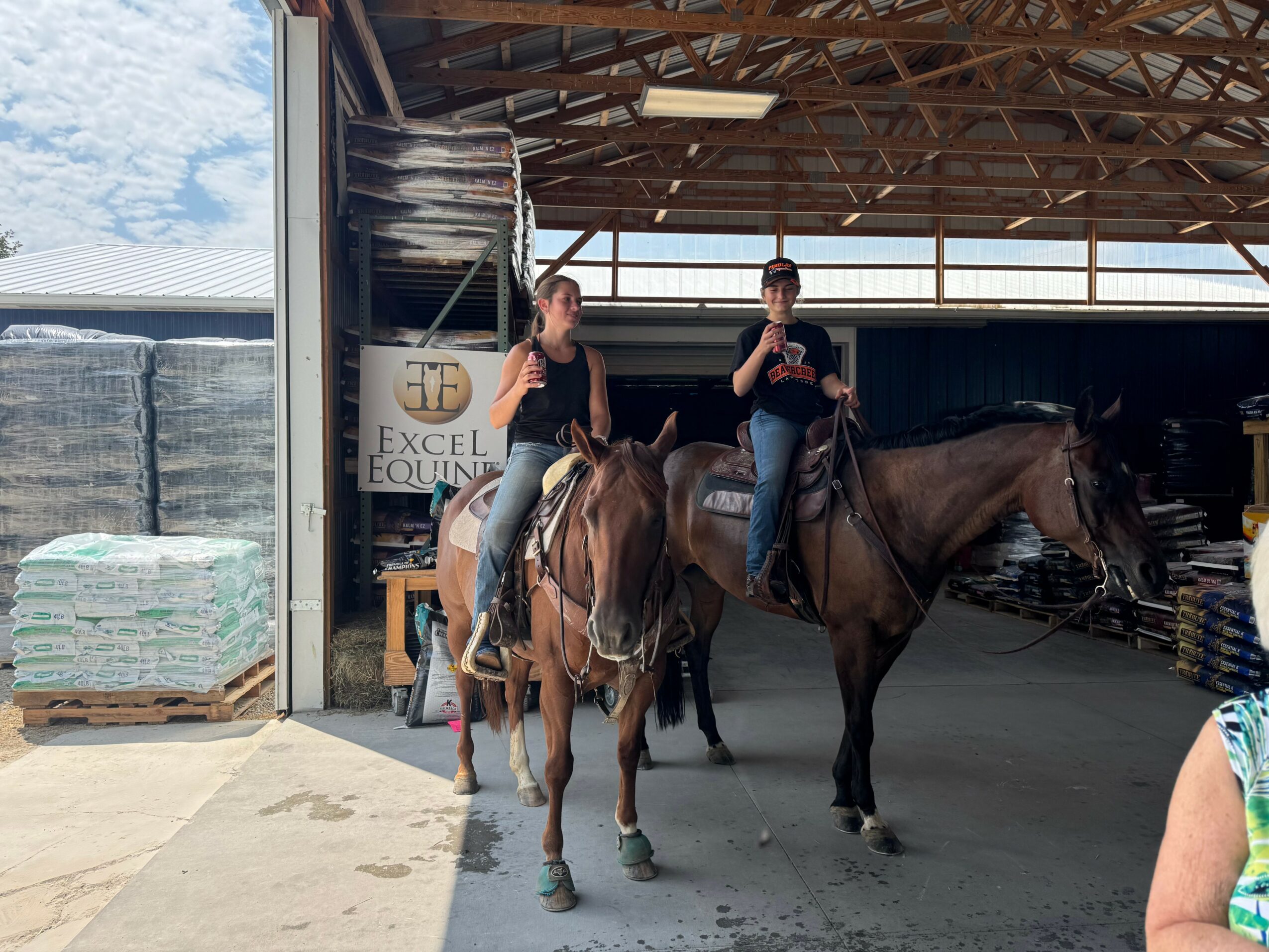 Two people on horseback inside a barn, with a sign reading 'Excel Equine' in the background.