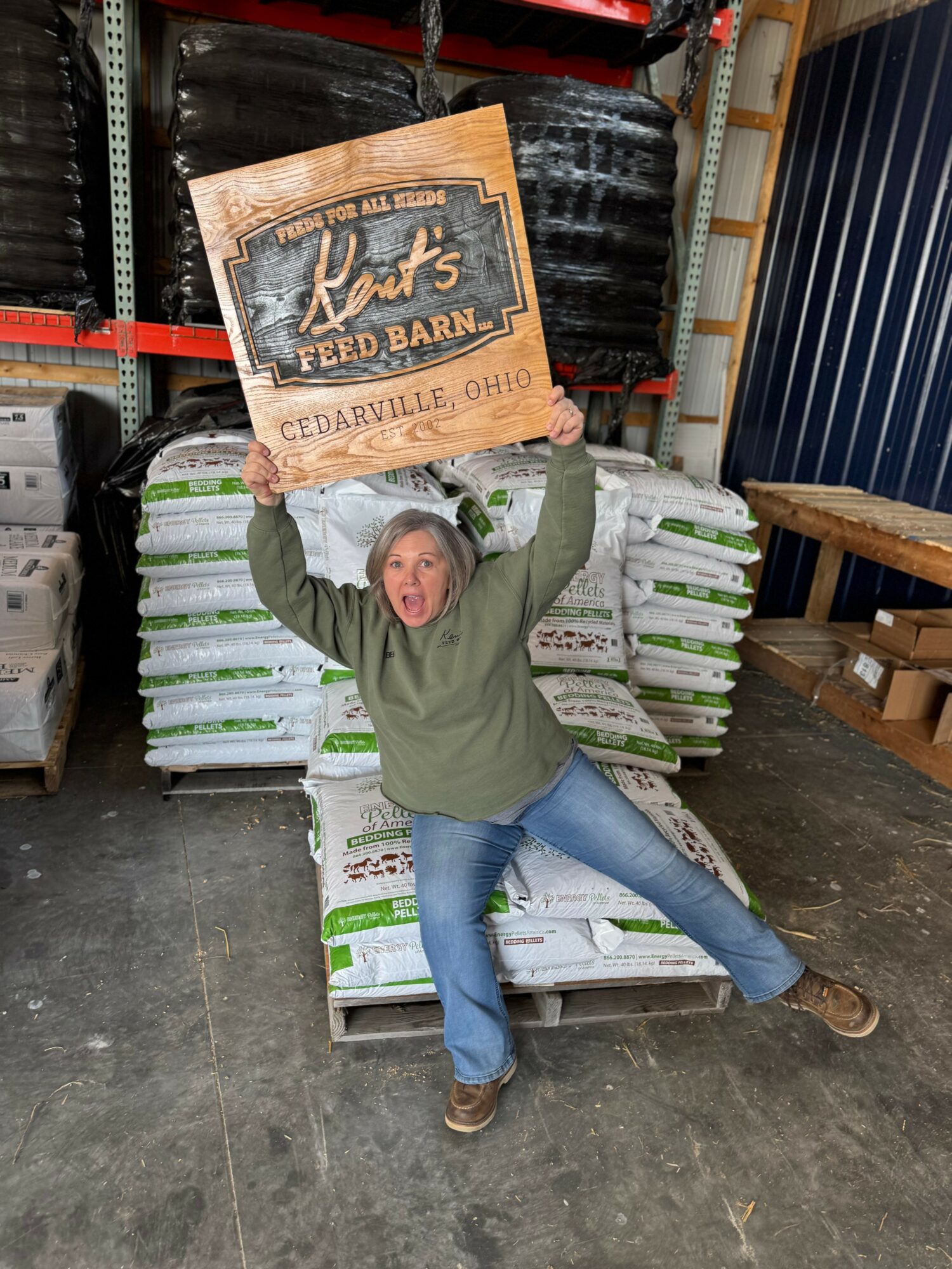 Person holding a sign above their head in a warehouse, surrounded by bags of soil or fertilizer, smiling.