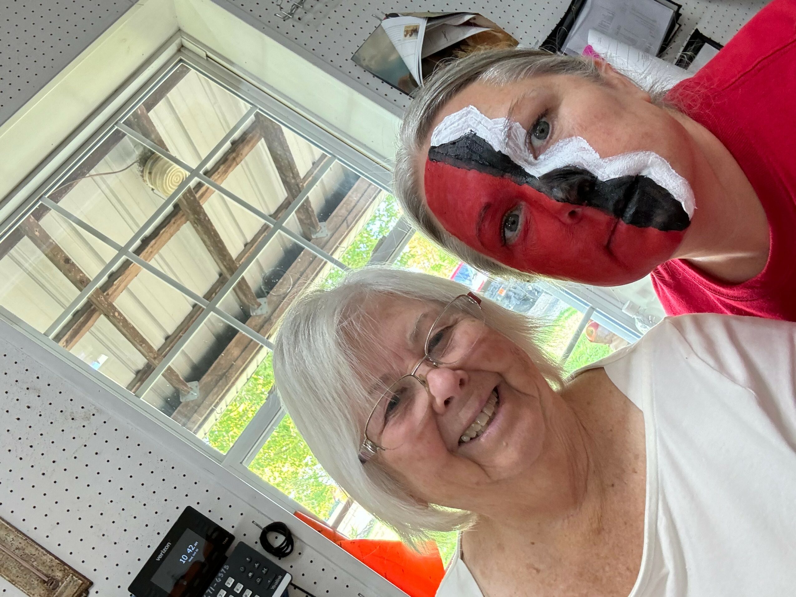 Two women smiling indoors near a window, one with face paint, the other with glasses and white hair.