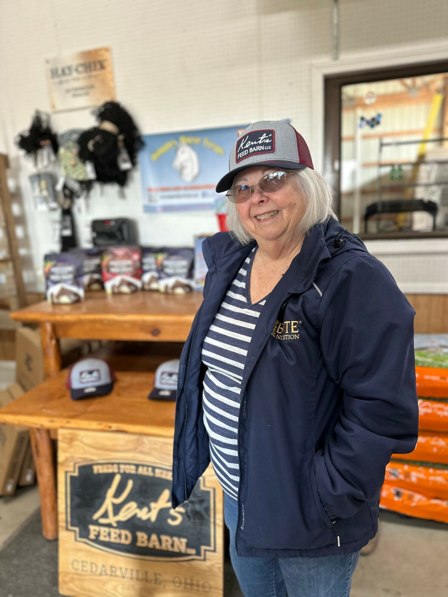 Smiling woman with glasses and a cap, wearing a striped shirt and navy jacket, standing indoors near a display table.