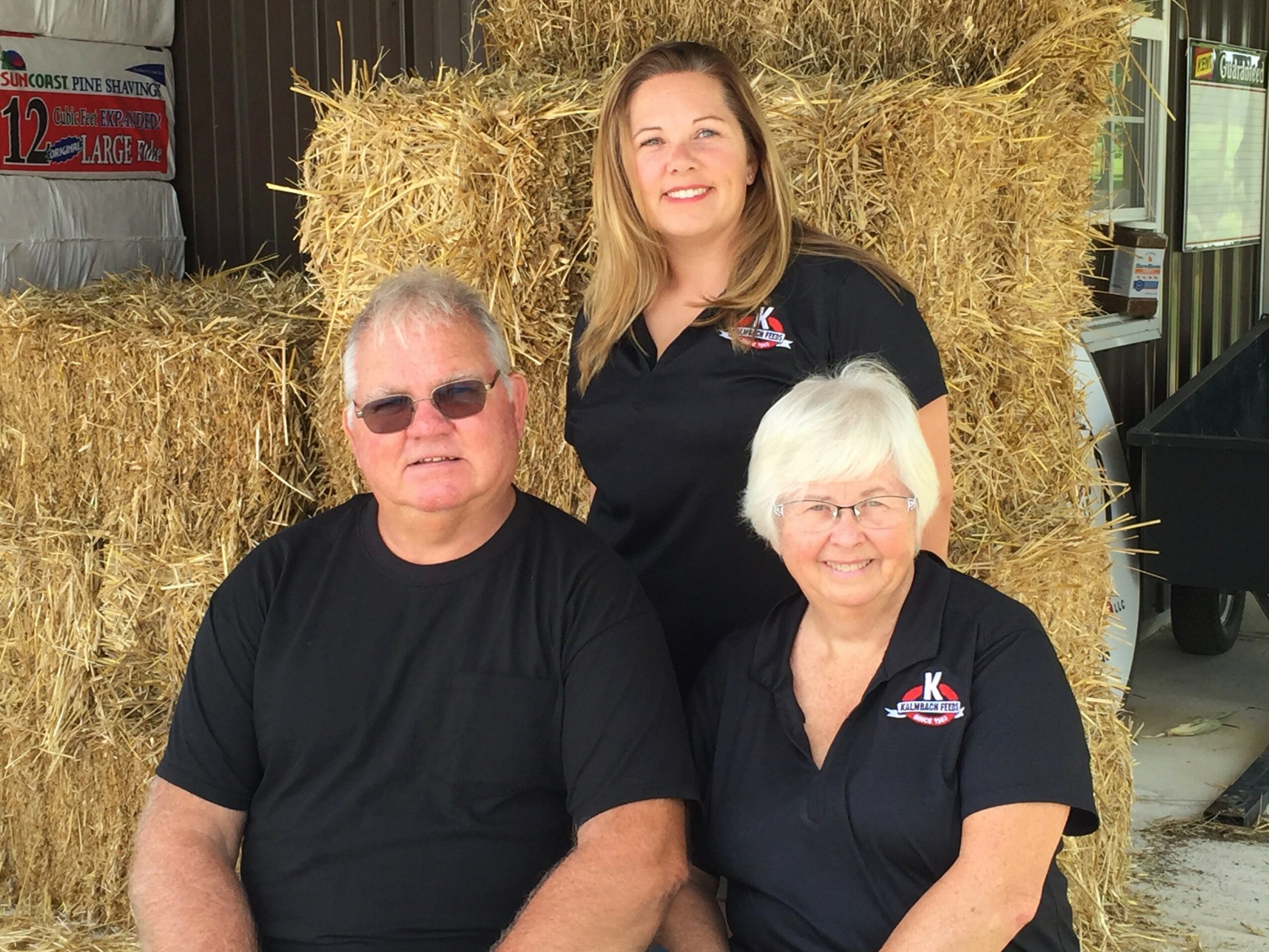 Two women and one man standing outdoors in front of hay bales, smiling, wearing black shirts with logos.
